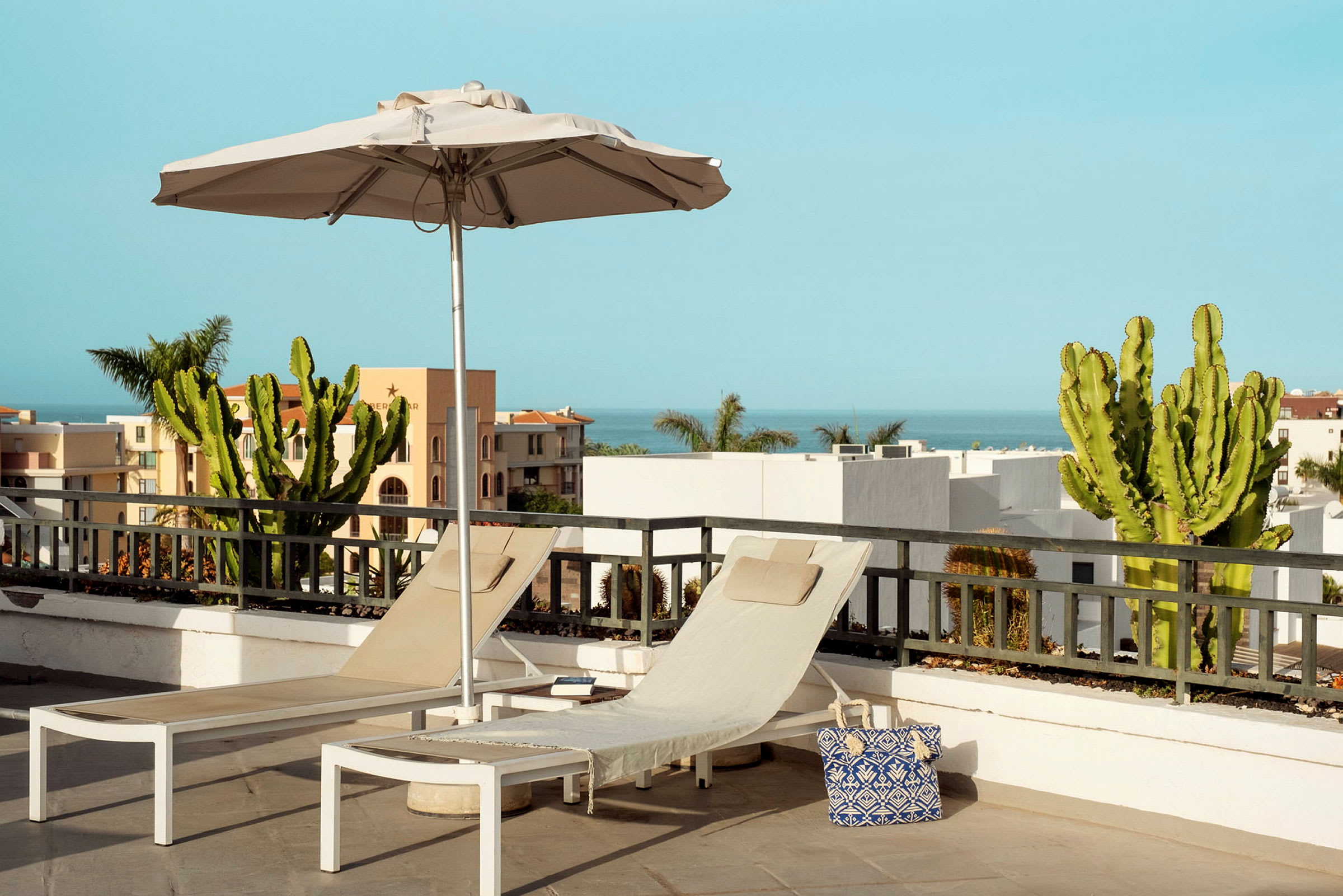 To liggestole og en parasol på en terrasse med kaktusser og udsigt mod havet. (AI-genereret tekst)