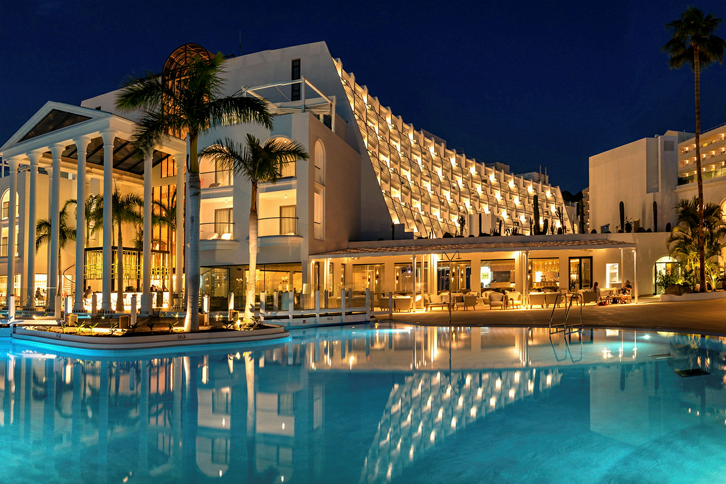 Hotel om natten med oplyst facade og swimmingpool i forgrunden. (AI-genereret tekst)