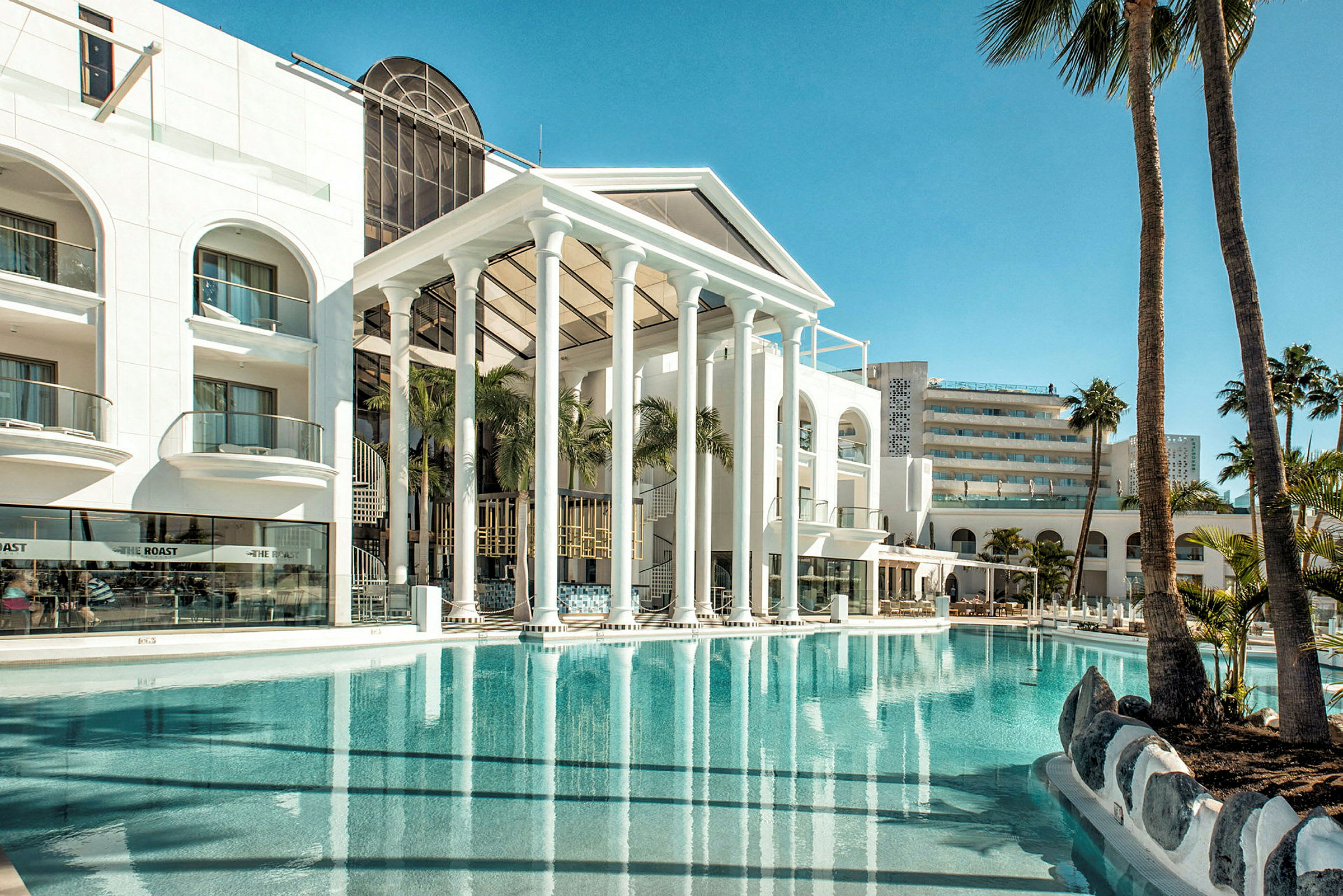 Hotelfacade med pool og palmer i forgrunden. (AI-genereret tekst)
