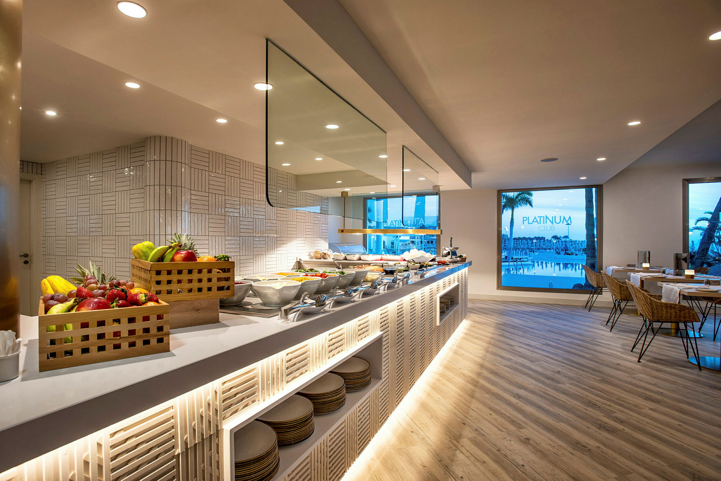 Billede af en hotelbuffet med frugtkurve, madopstillinger og siddepladser nær store glasvinduer. (AI-genereret tekst)