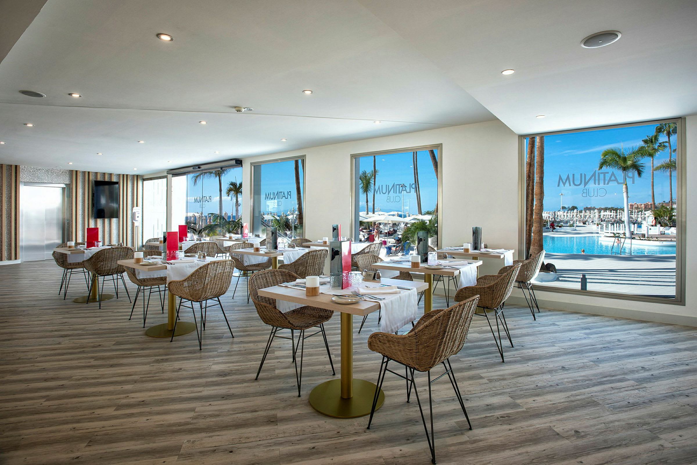 Indretning af en lys restaurant med udsigt til en pool og palmer gennem store vinduer. (AI-genereret tekst)