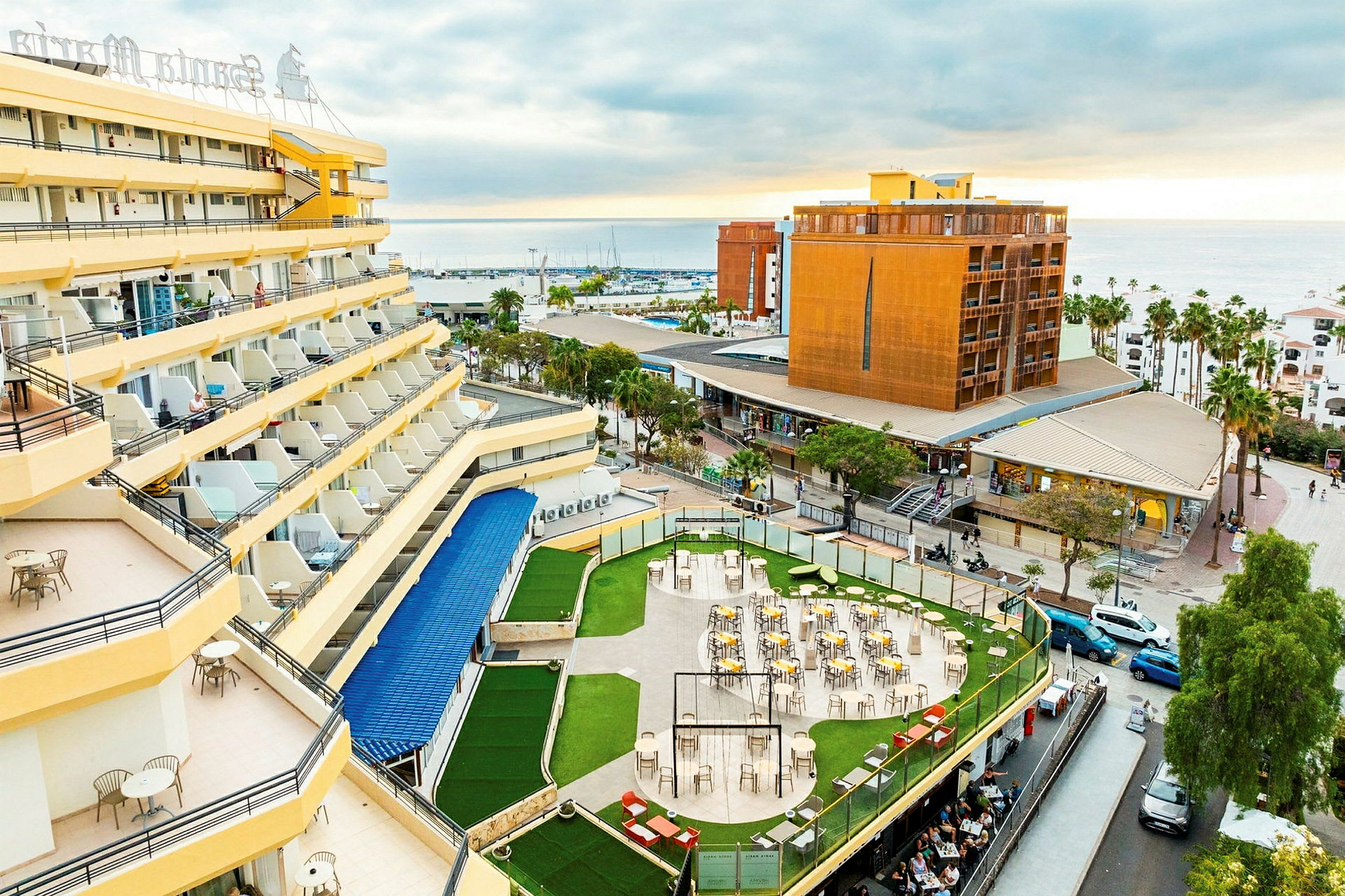 Hotel med gule facader og altaner, en terrasse med borde og stole omgivet af grønne områder samt en udsigt over byen og havet i baggrunden. (AI-genereret tekst)