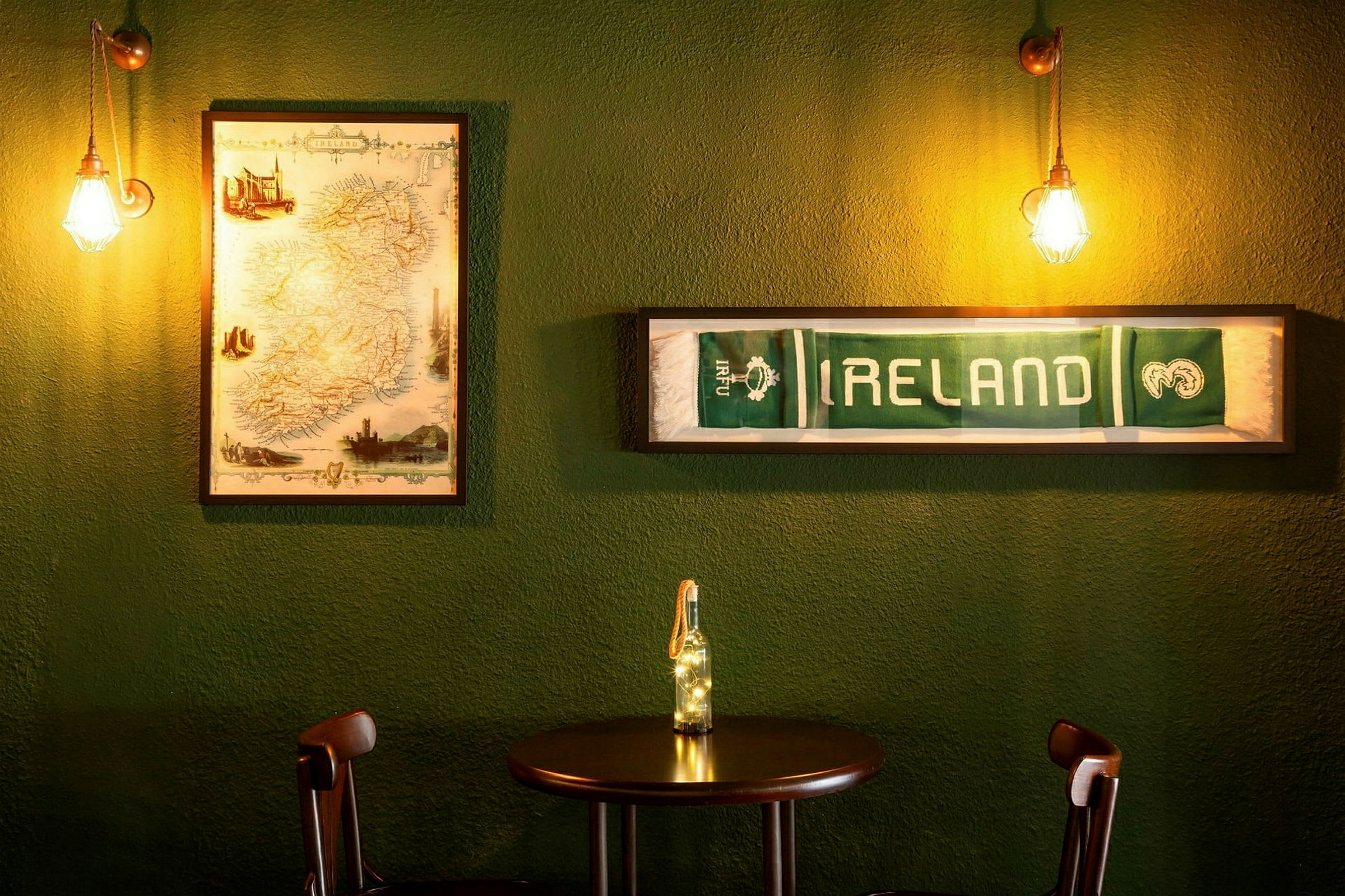 Et bord med to stole foran en grøn væg, dekoreret med et indrammet kort over Irland og et indrammet tørklæde med teksten "IRELAND". (AI-genereret tekst)