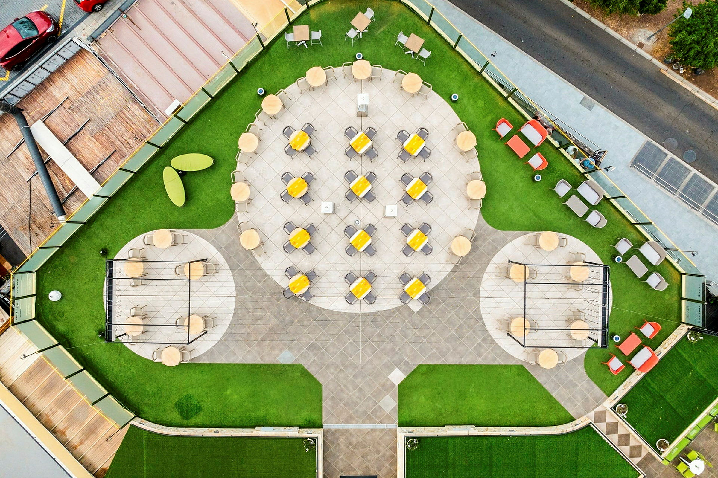 Luftfoto af en haveagtig udendørs terrasse med borde, stole og parasoller grupperet på en cementeret overflade, omgivet af grønt græs. (AI-genereret tekst)