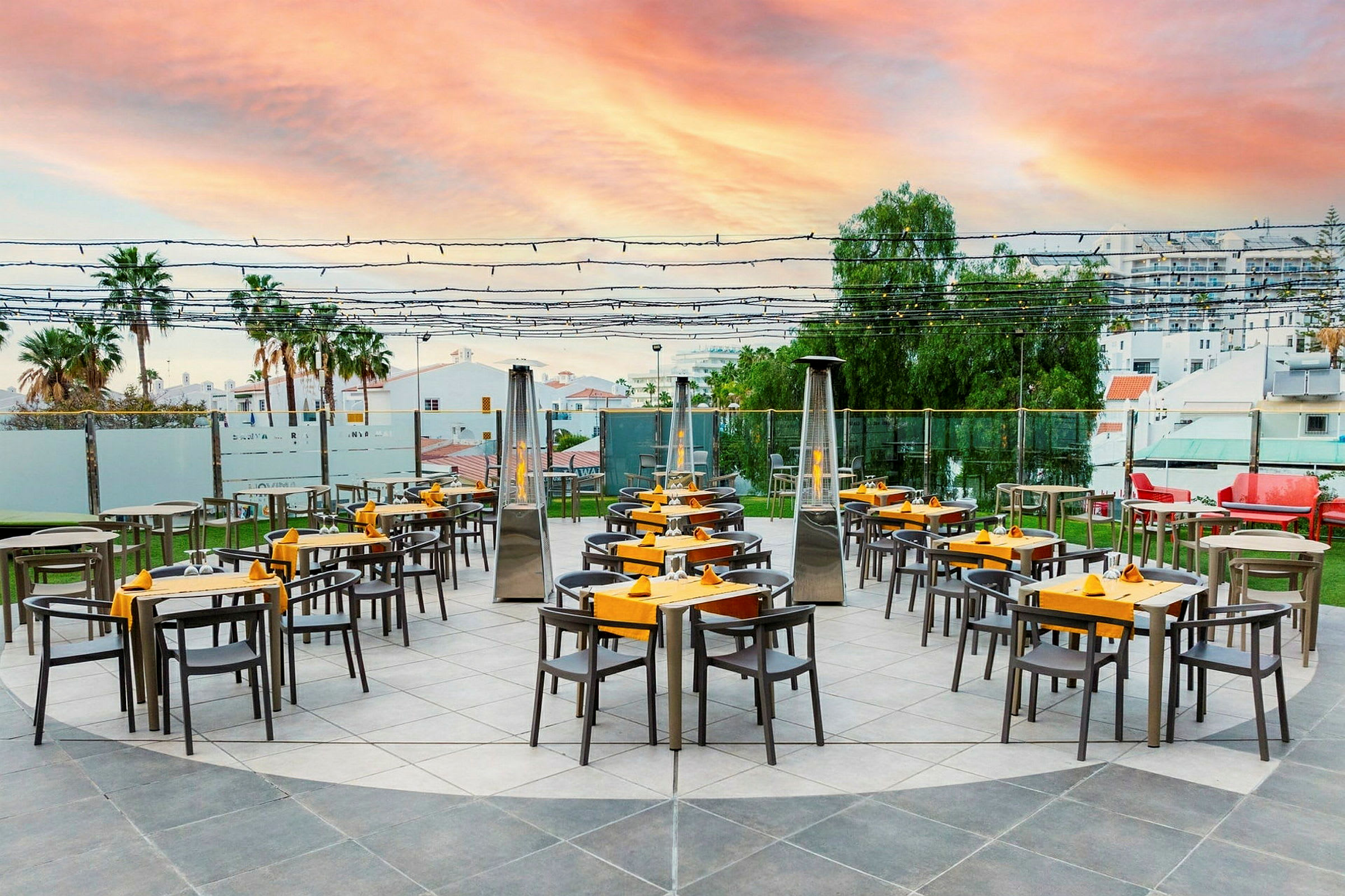 En udendørsrestaurant med borde og stole arrangeret på en brolagt flade under en himmel med solnedgangens farver. (AI-genereret tekst)