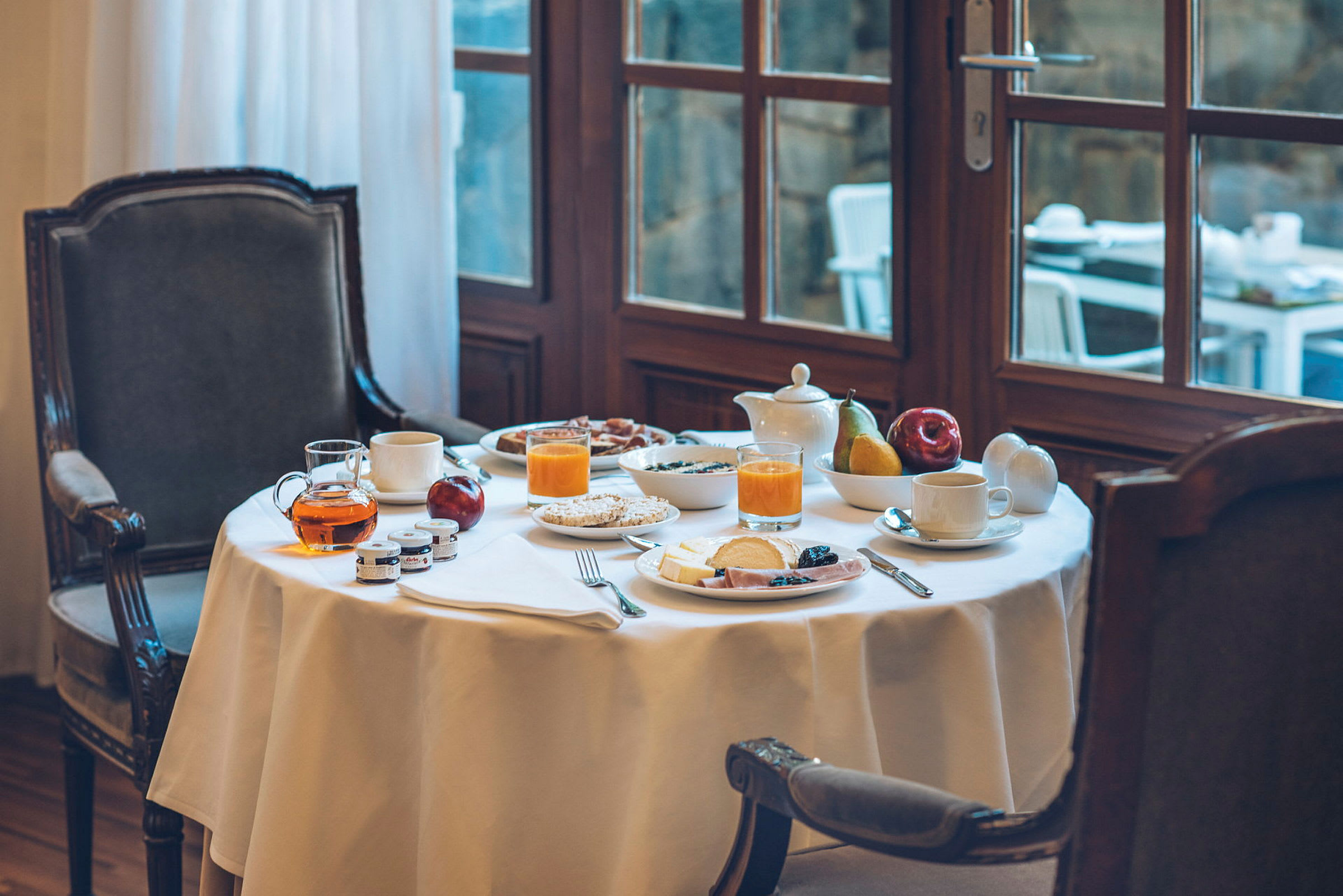 Et dækket morgenmadsbord med glas appelsinjuice, te, frugt og skiveskåret pålæg. Bordet står ved et vindue. (AI-genereret tekst)