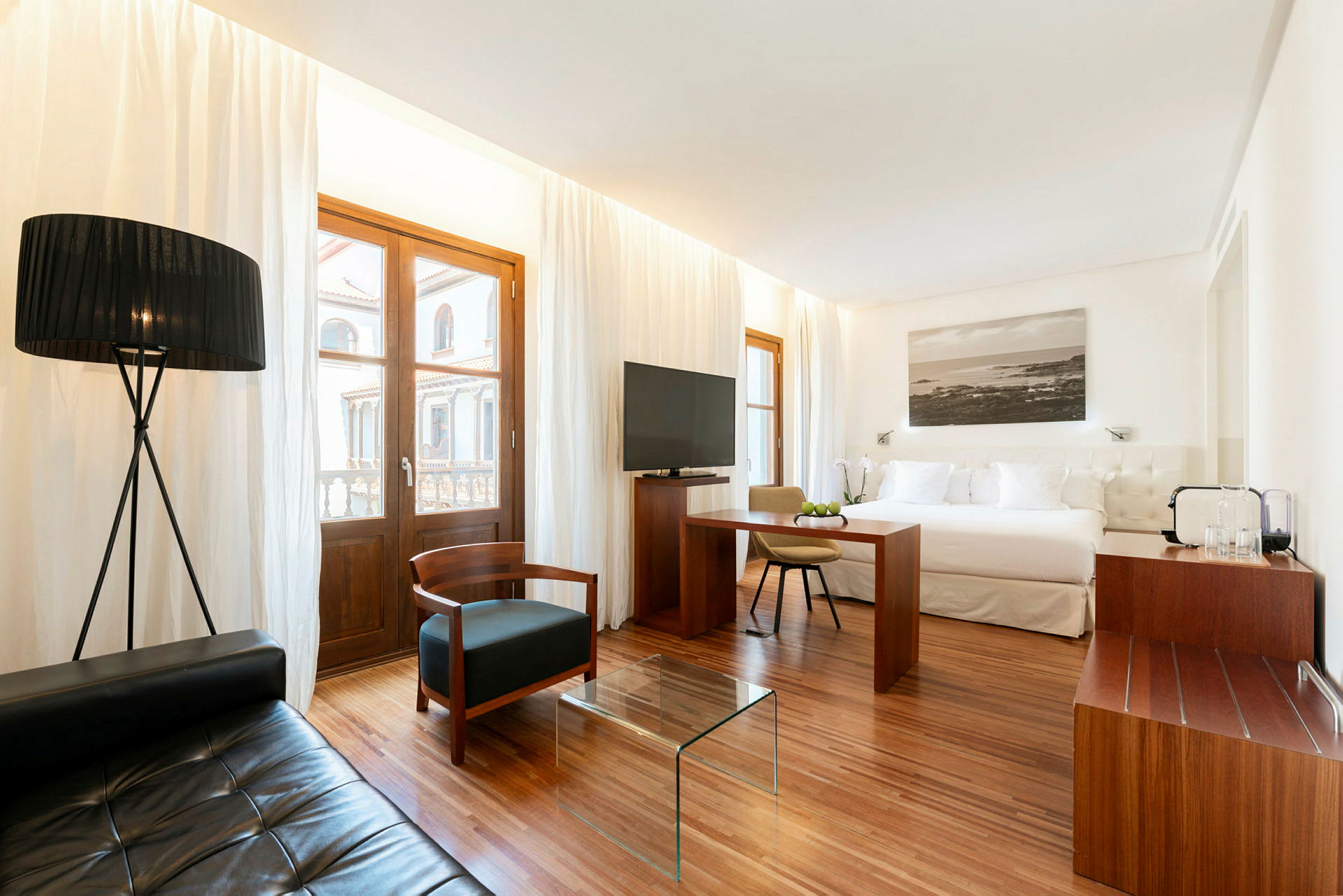 Et moderne indrettet hotelværelse med en stor seng, siddeområde med tekstilmøbler og et glasbord, samt vinduer med hvide gardiner. (AI-genereret tekst)