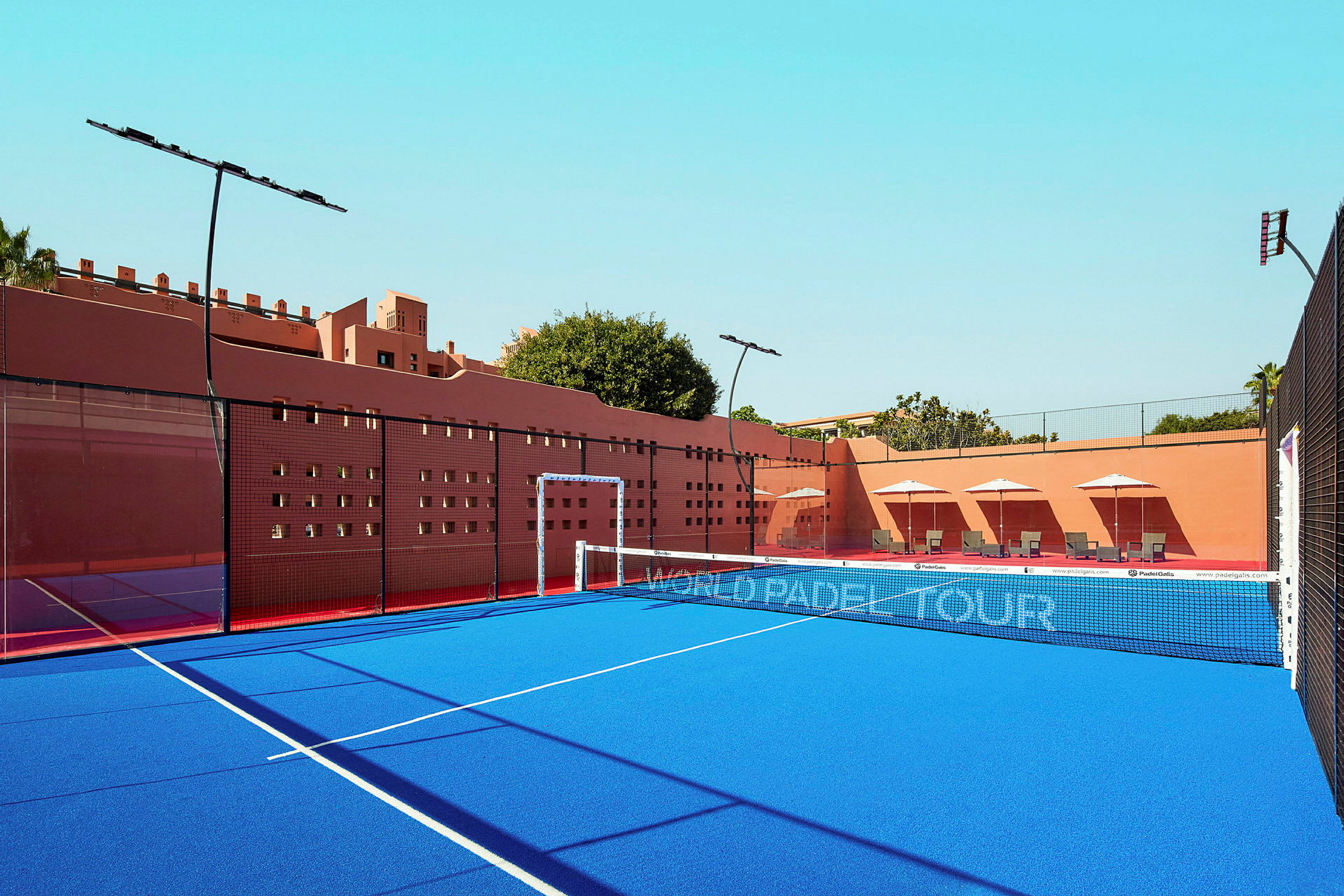Padelbane med blåt underlag omgivet af lyserøde vægge. Liggestole med parasoller ses i baggrunden. (AI-genereret tekst)