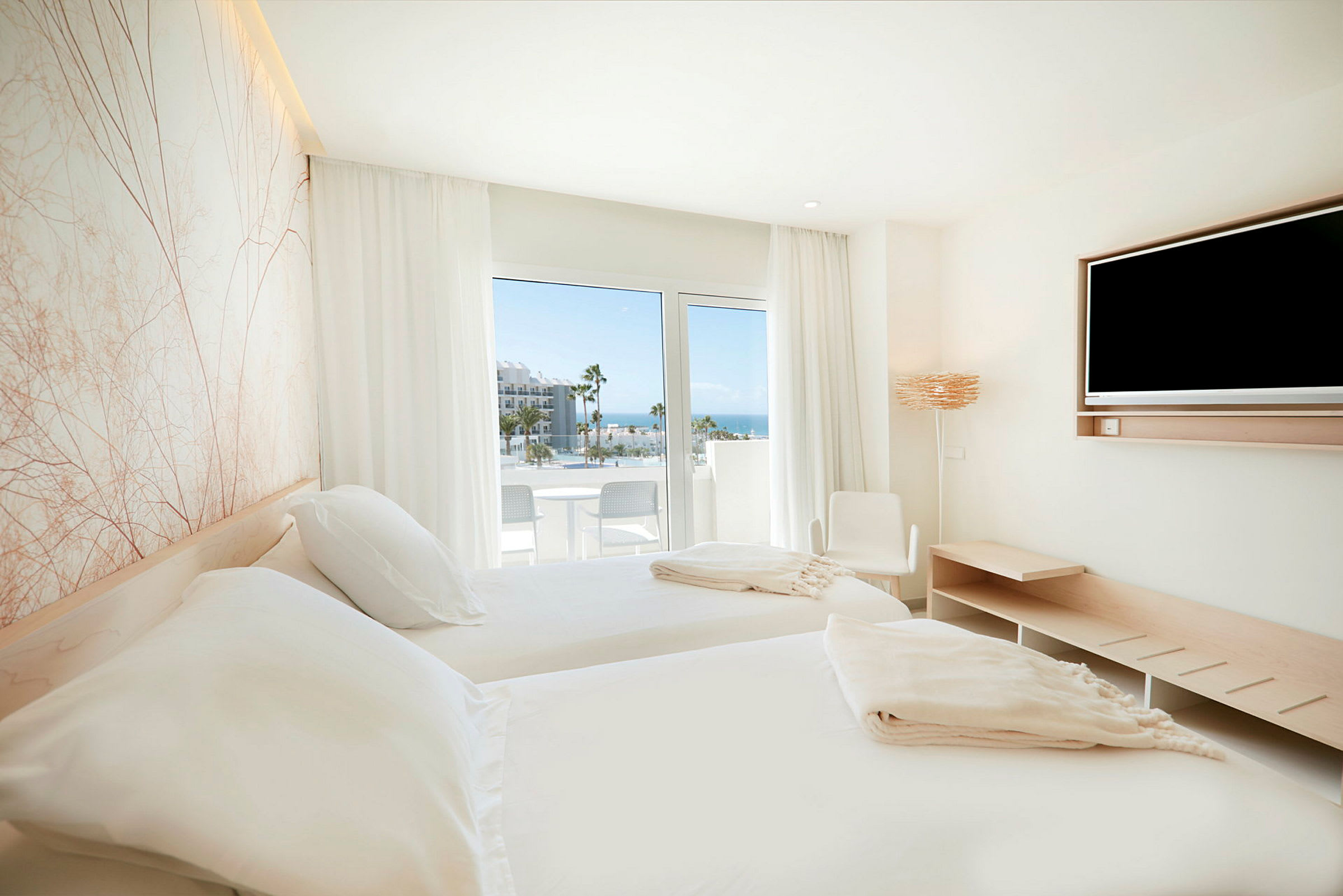 Et lyst soveværelse med to enkeltsenge, et TV og en balkon med udsigt over havet. (AI-genereret tekst)