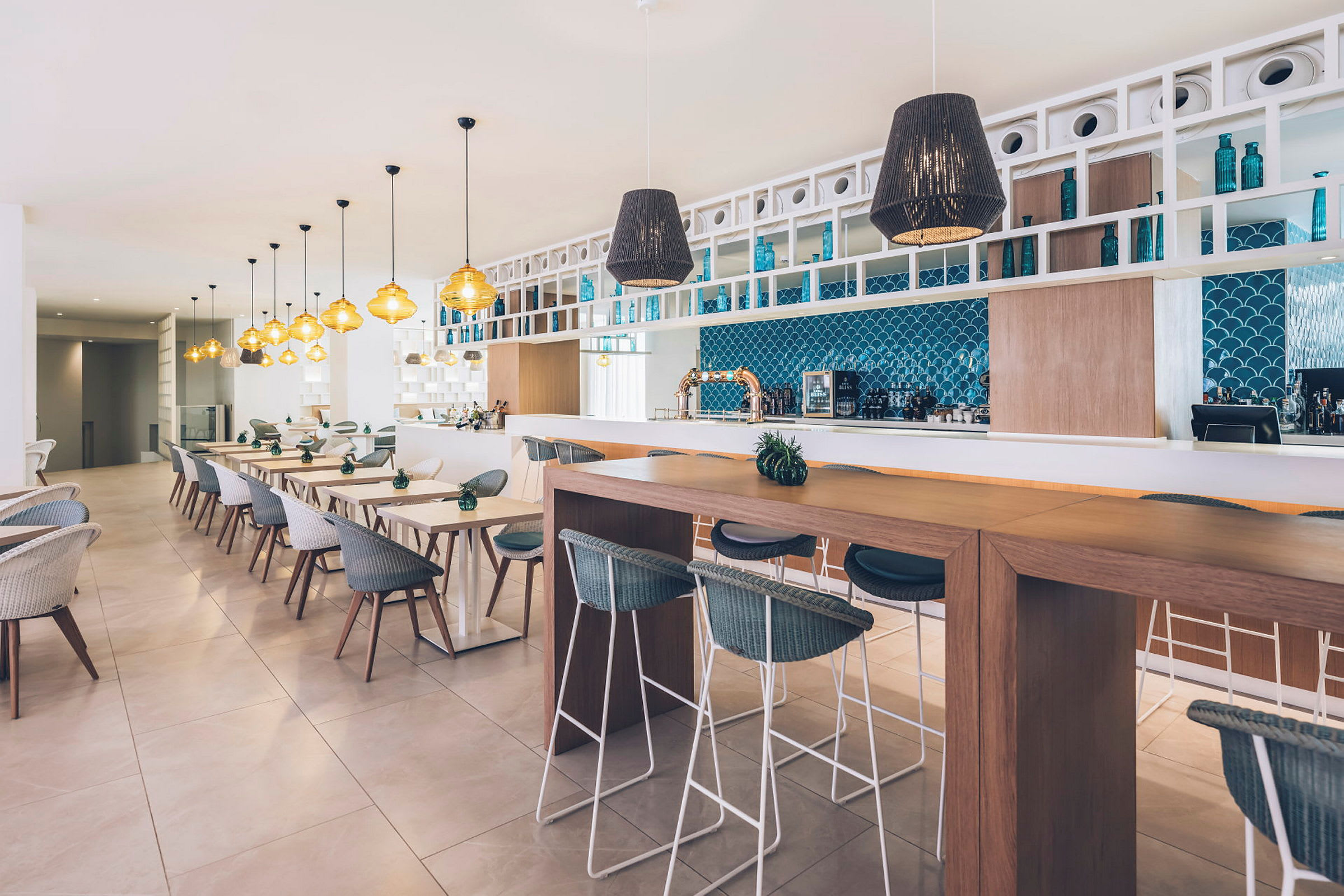 Billede af en moderne og stilfuld restaurant med borde og stole, varme belysningsdetaljer og en væg med blå mosaik ved baren. (AI-genereret tekst)