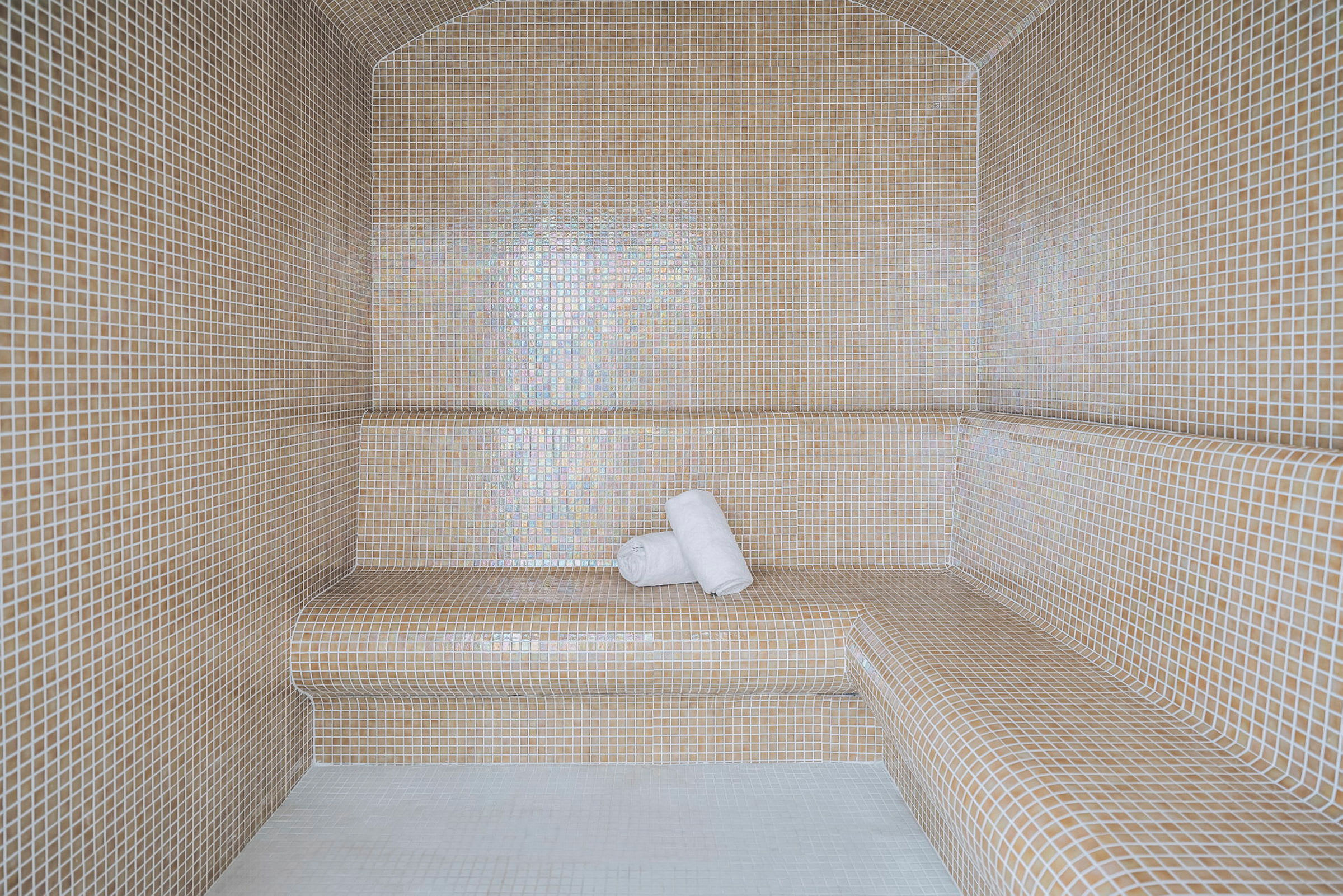 Et dampbad med vægge og bænke beklædt med små beige mosaikfliser. To hvide håndklæder ligger på en af bænkene. (AI-genereret tekst)