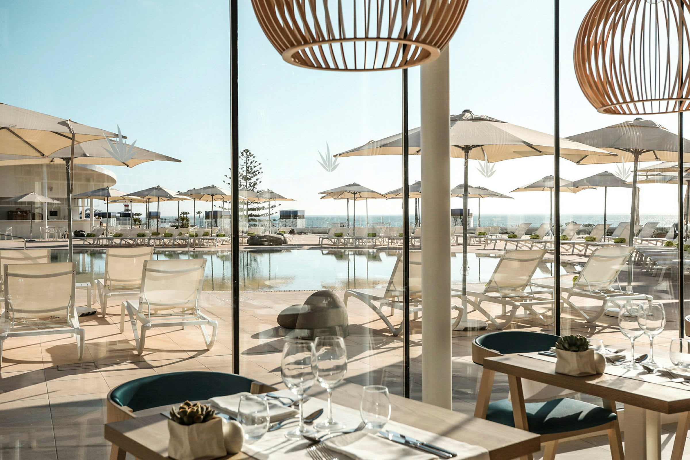 Udsigt fra en restaurant med borde og glasopdækning mod en terrasse med liggestole og parasoller ved en pool. (AI-genereret tekst)
