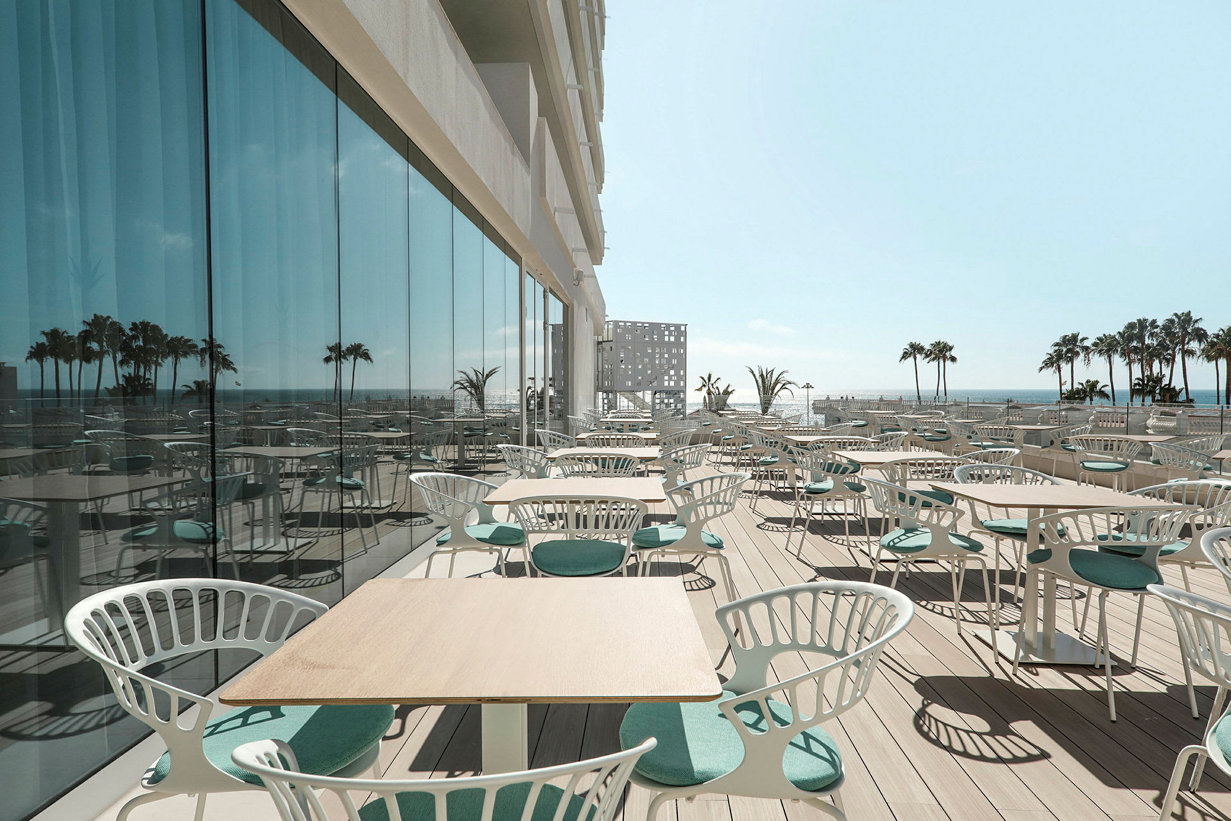 En udendørs terrasse med borde og stole med udsigt over havet og palmer i det fjerne. Solskinsvejr. (AI-genereret tekst)