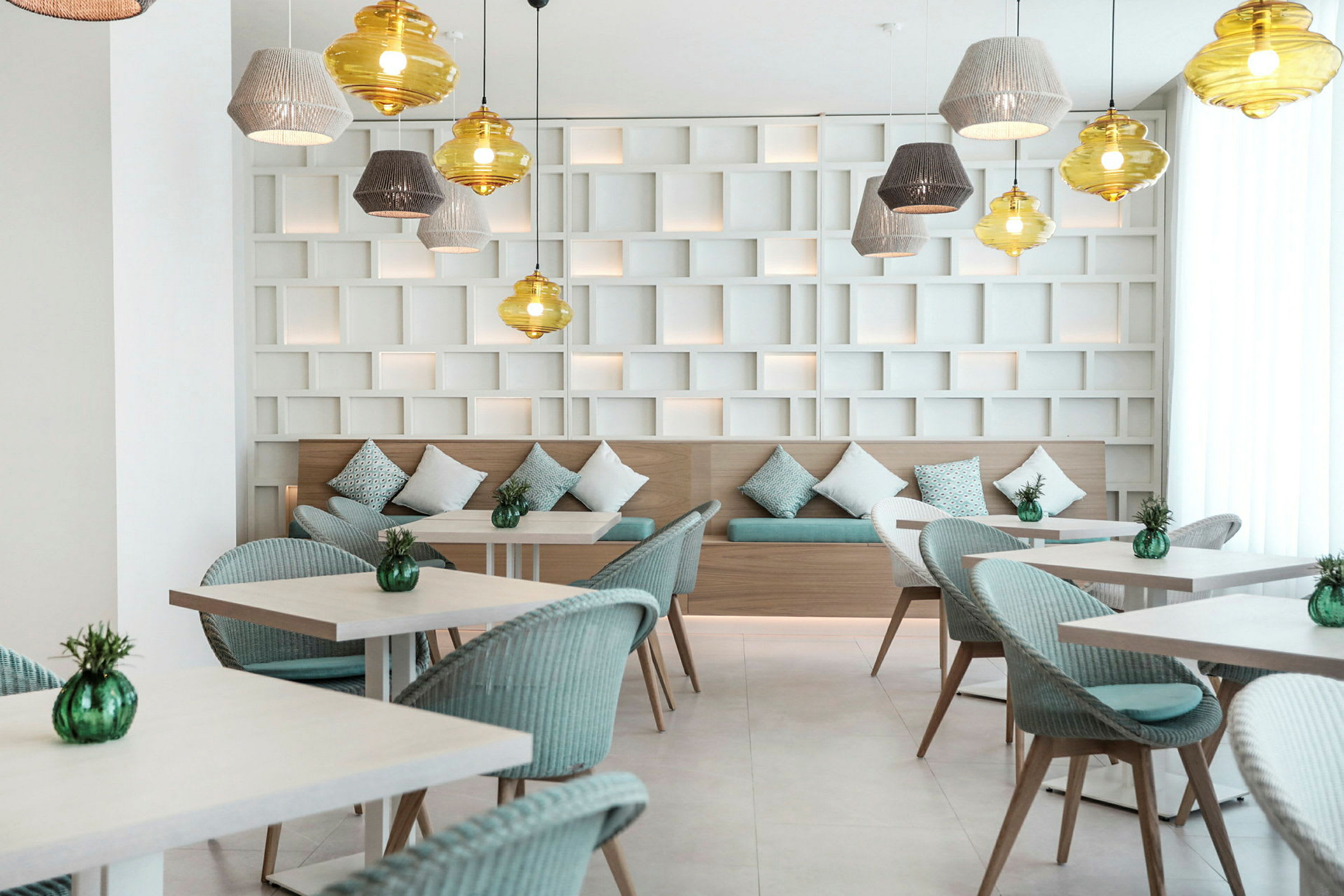 Et moderne caférum med borde, stole og dekorative lamper i loftet. Bagvæggen har et design med geometriske former, og bænken har puder. Flasker med grønne blade står på bordene. (AI-genereret tekst)