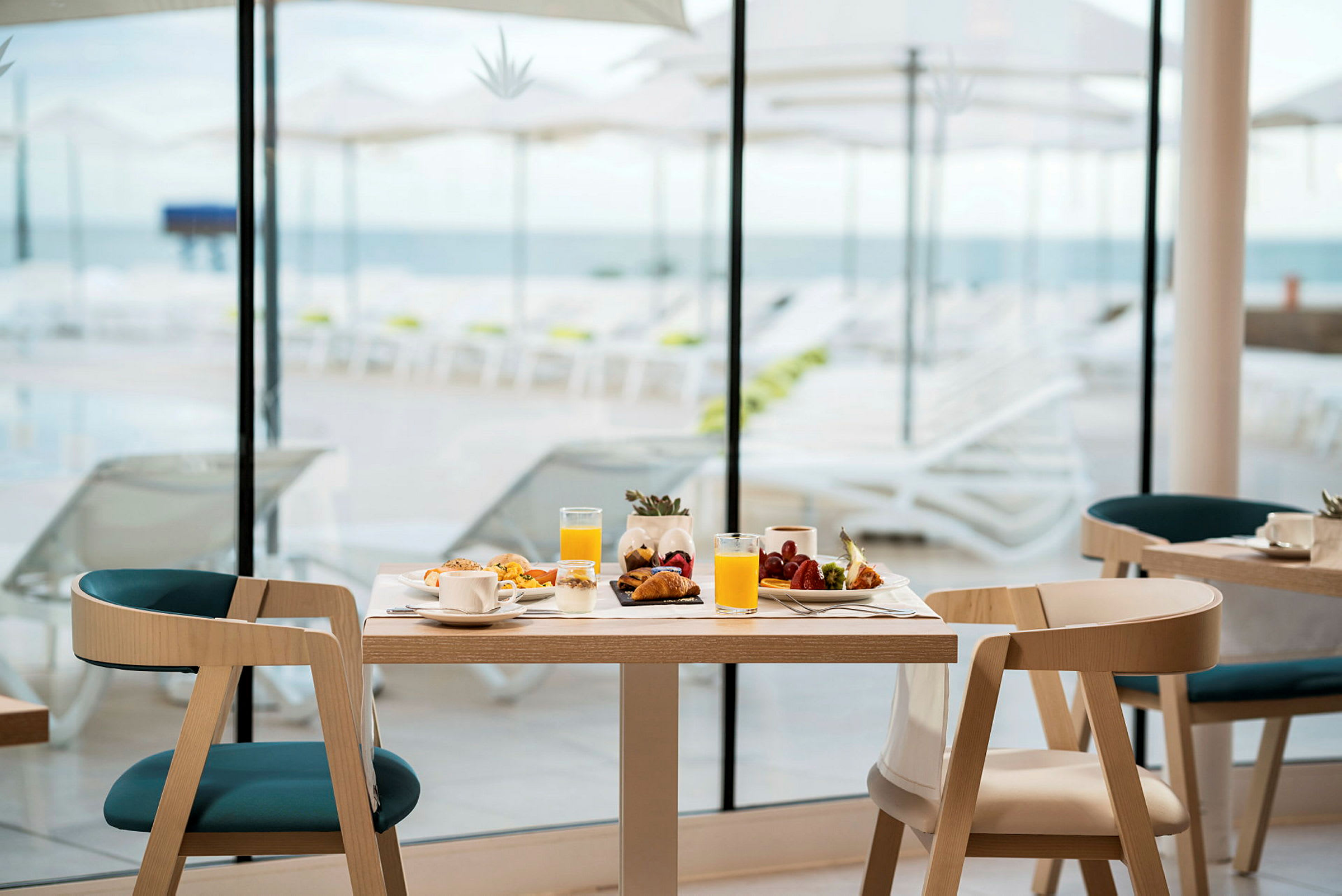 Et dækket bord med morgenmadsmad og drikke ved siden af store vinduer, der vender ud mod en solrig terrasse med liggestole og havudsigt. (AI-genereret tekst)