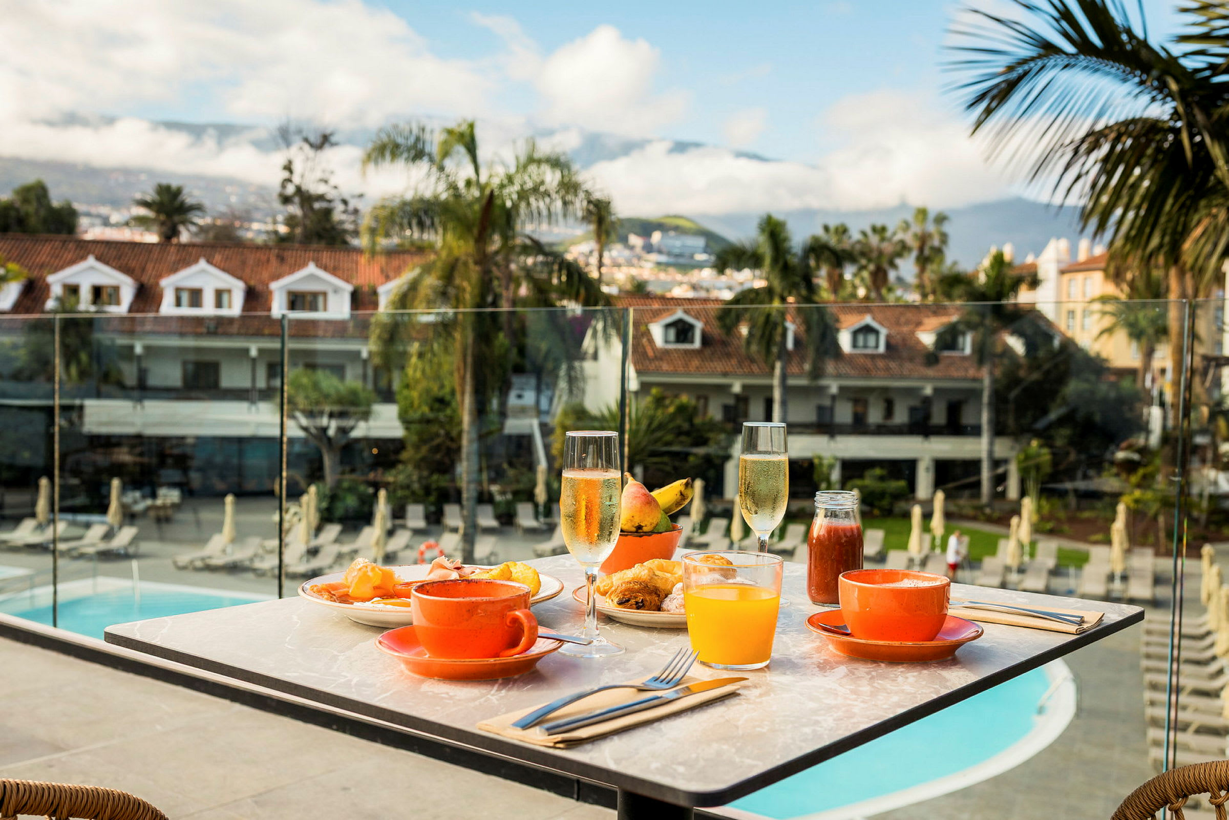 Et dækket bord med morgenmad og drikkevarer på en terrasse med udsigt over et poolområde og de omkringliggende omgivelser. (AI-genereret tekst)