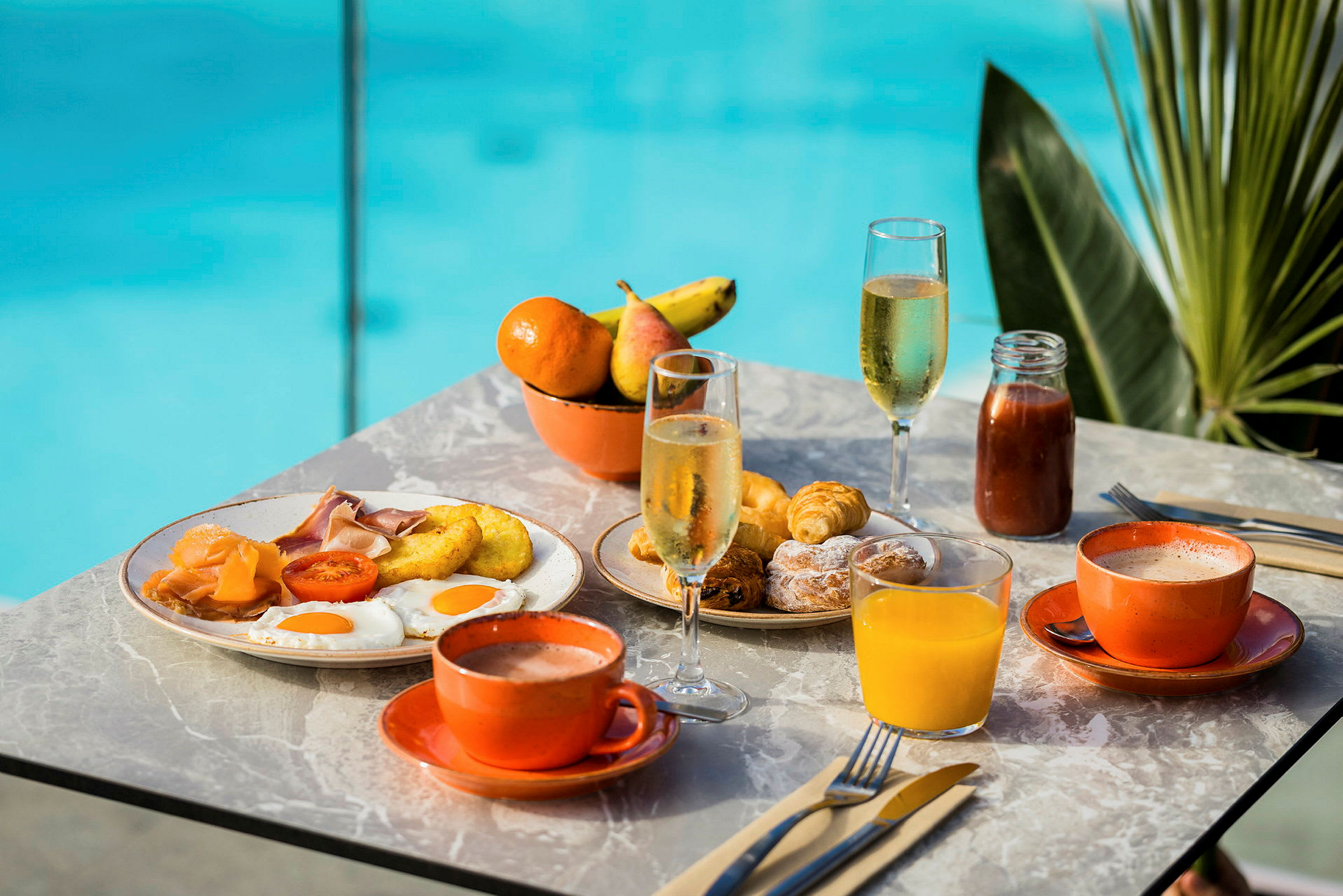 Morgenbord med forskellige retter som æg, pølser, croissanter og drikkevarer som juice og kaffe. En pool ses i baggrunden. (AI-genereret tekst)