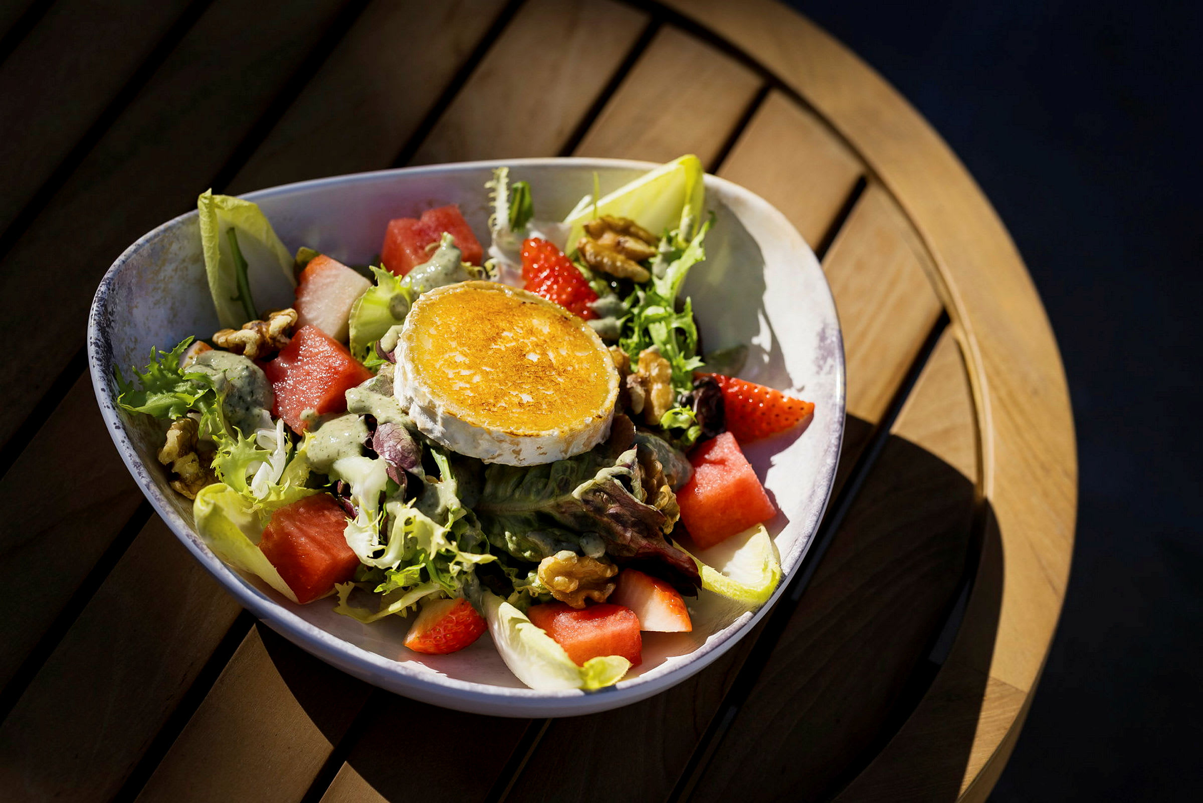 En tallerken med en salat, der indeholder grillet osteskive, melon, jordbær, nødder og salatblade. Tallerkenen står på et træbord. (AI-genereret tekst)