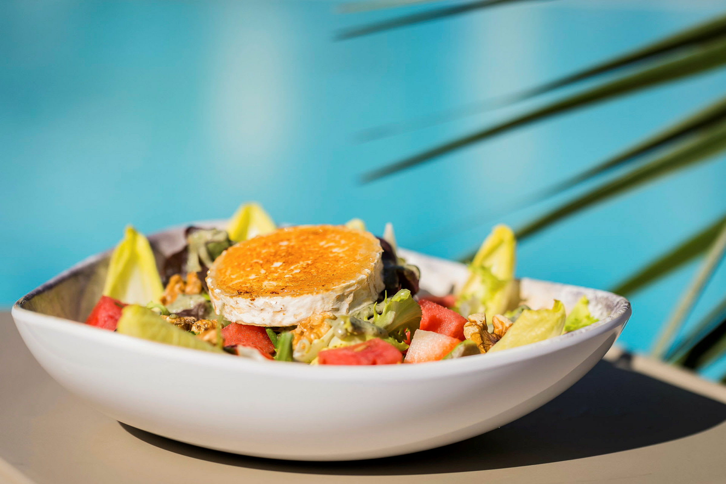 En salat med forskellige grøntsager og et stykke gratineret ost, serveret i en hvid skål med sløret baggrund af blåt vand og grønne blade. (AI-genereret tekst)