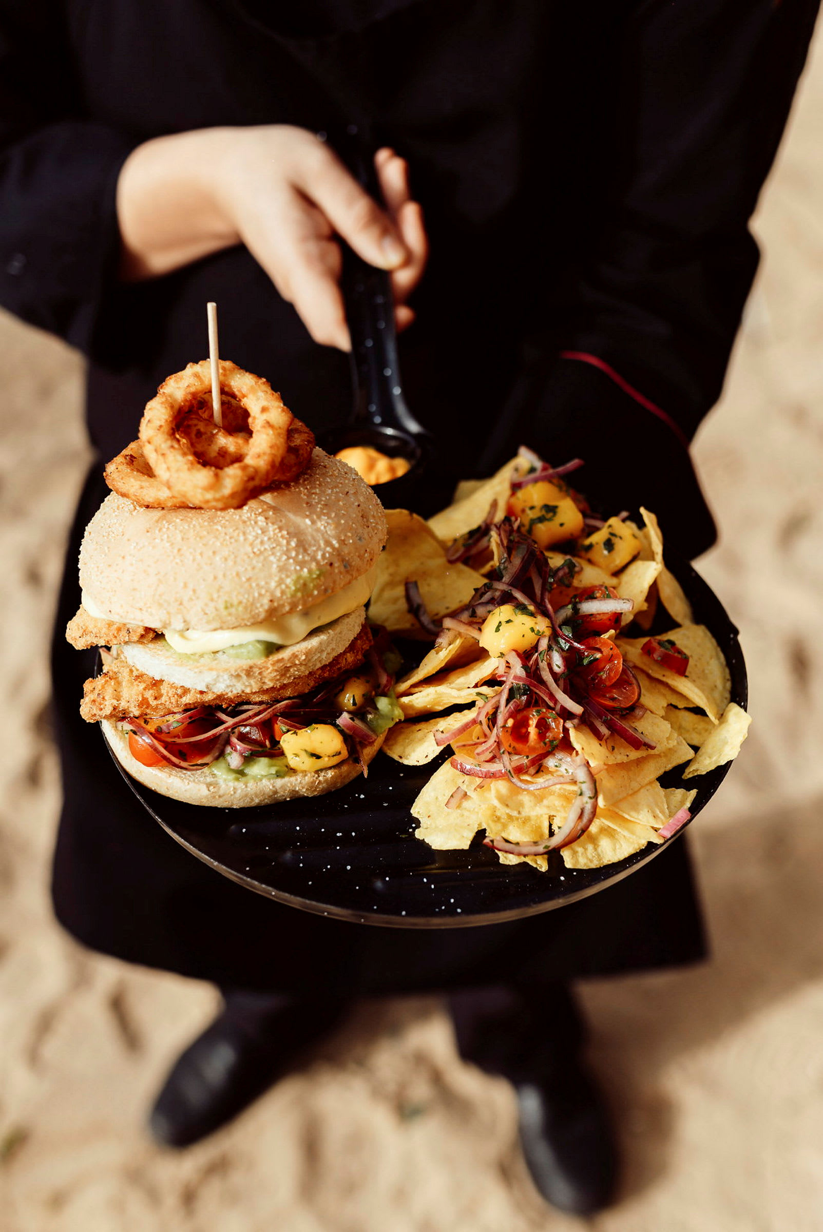En person holder en tallerken med en burger toppet med løgringe og en side af nachos med salat. (AI-genereret tekst)