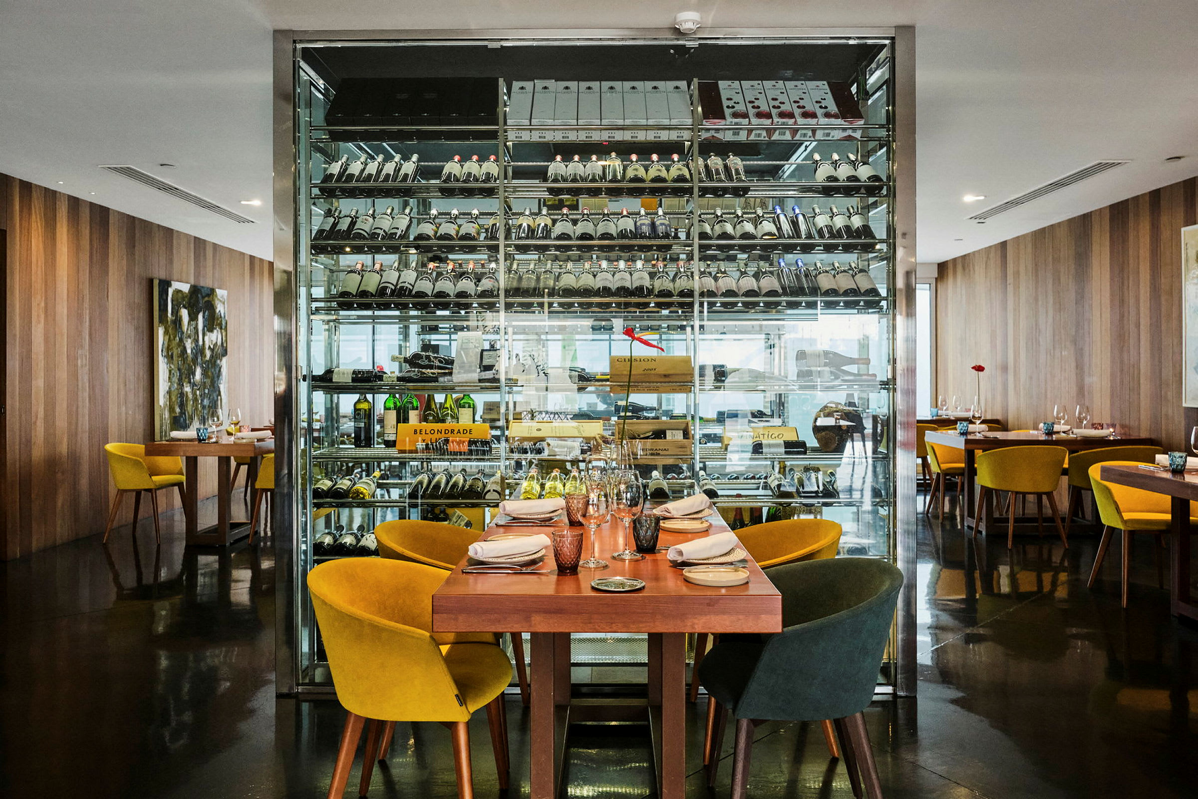 Restaurant med et bord klart til gæster foran en glasvæg med lagrede vinflasker. (AI-genereret tekst)