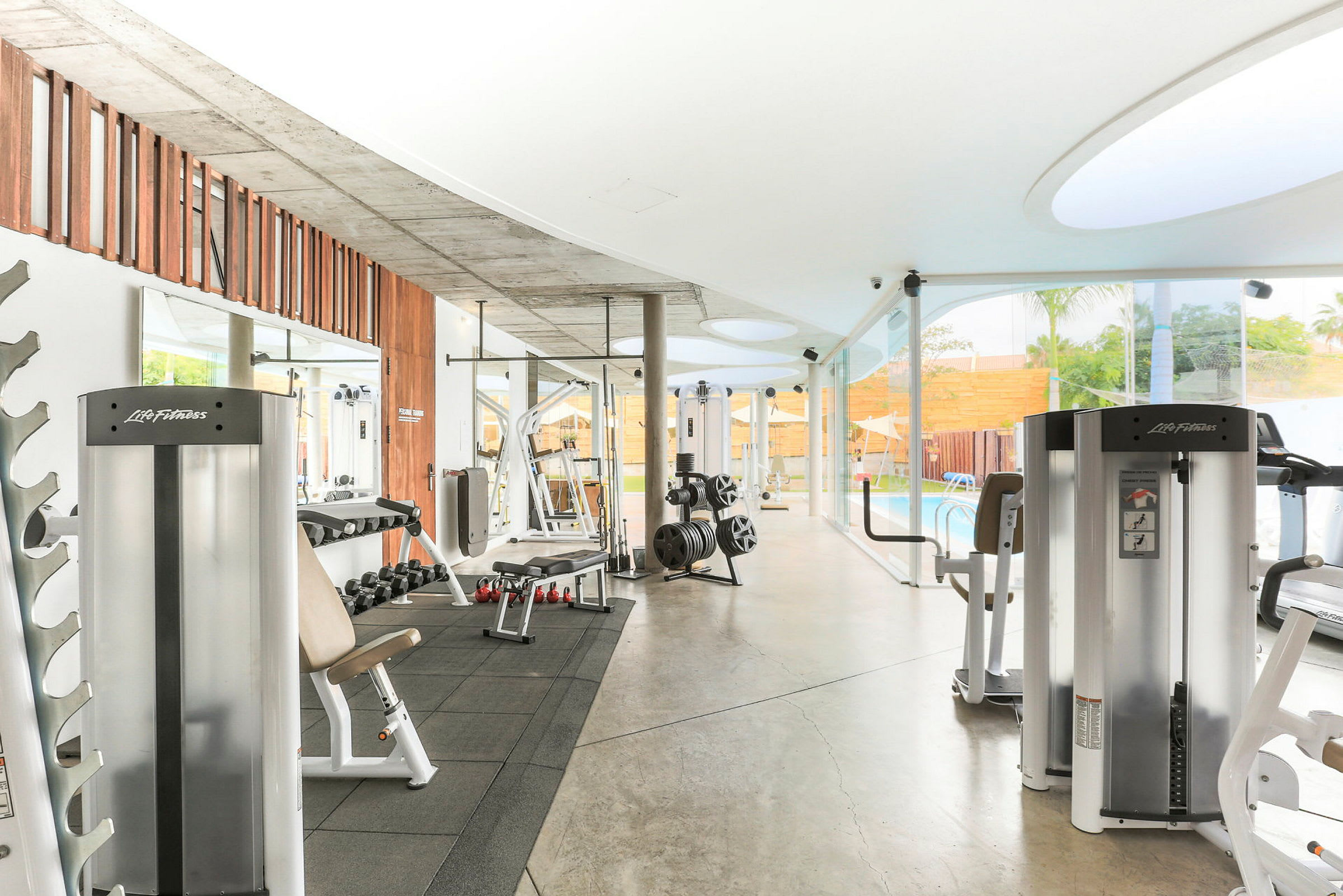 Et moderne fitnesscenter med træningsmaskiner og frie vægte, beliggende tæt på en glasvæg med udsigt til en pool. (AI-genereret tekst)