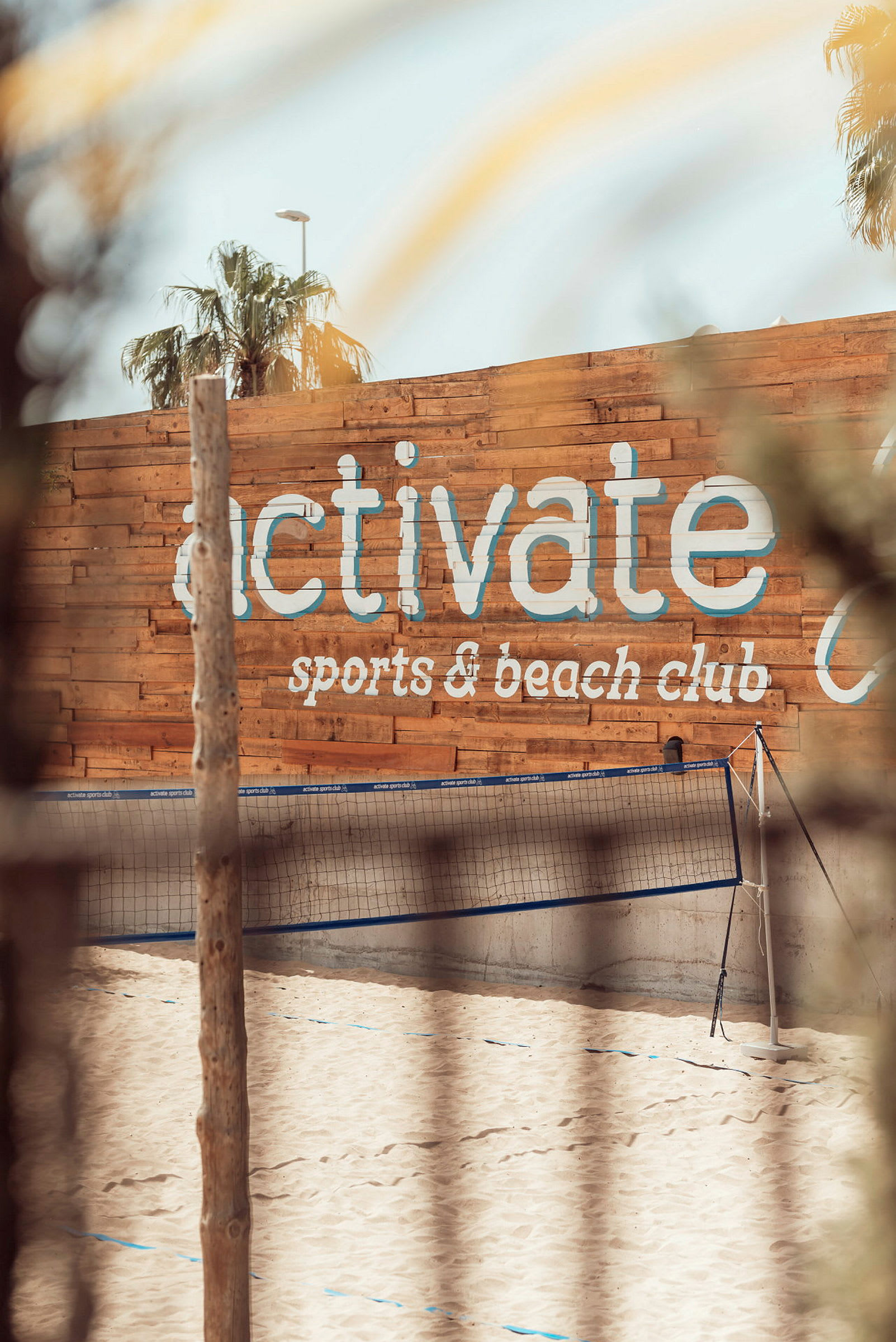 Sandstrand med volleyballnet foran en trævæg med teksten 'activate sports & beach club'. (AI-genereret tekst)