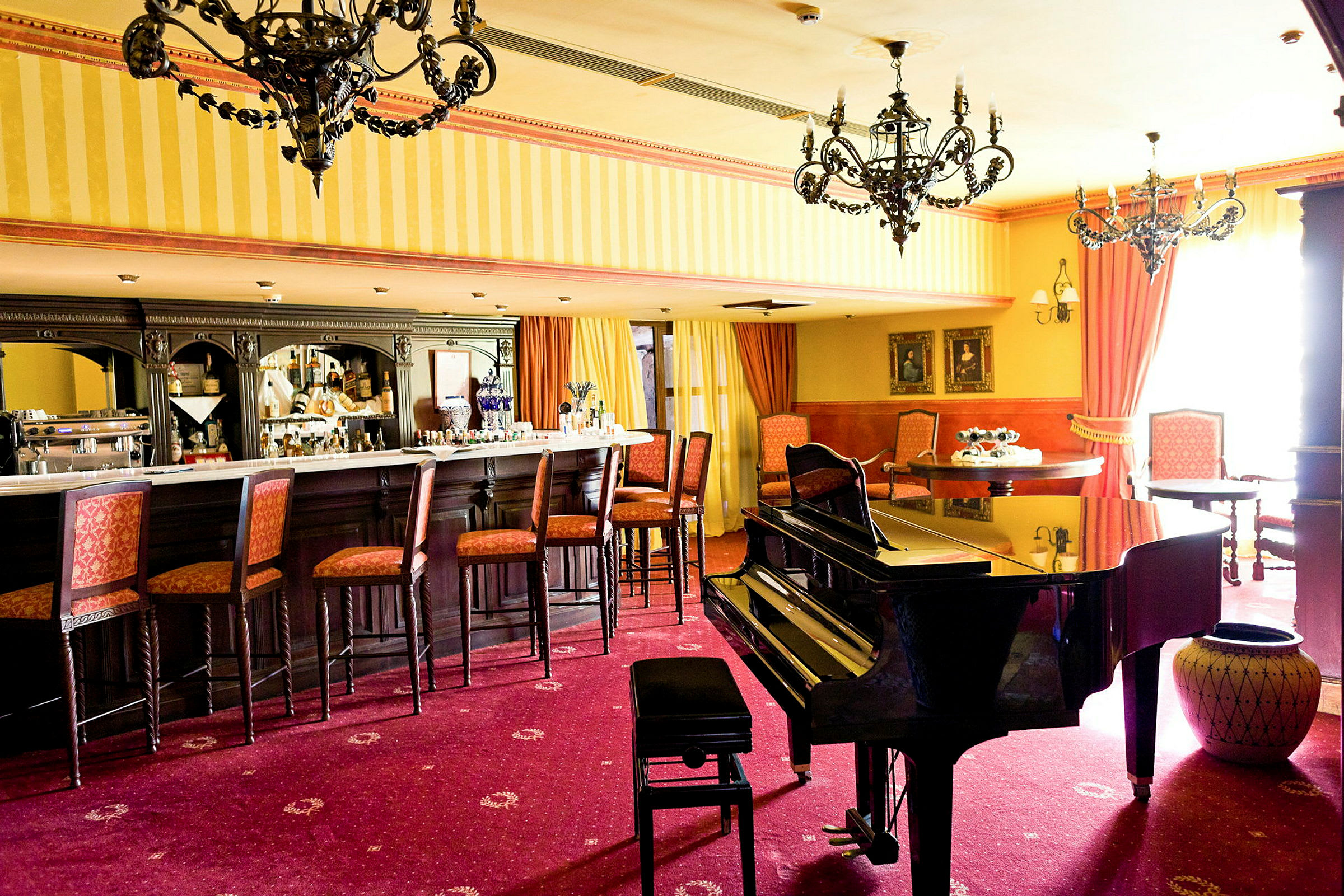 En barlounge med stole ved bardisken, et klaver i forgrunden og elegante lysekroner i loftet. (AI-genereret tekst)