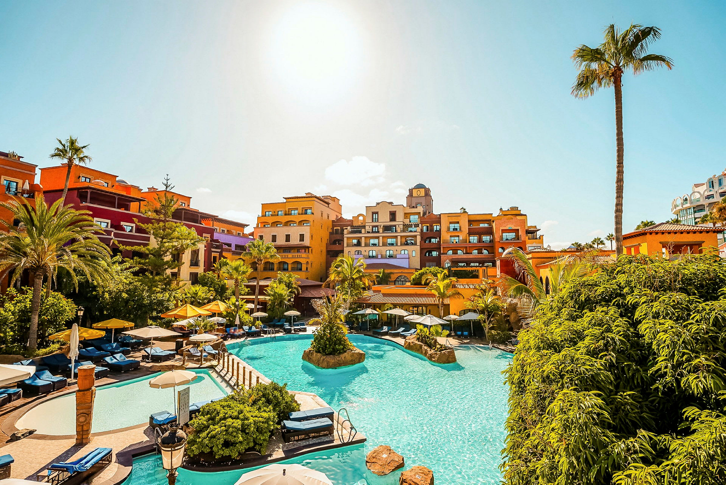 Ferieanlæg med pool, liggestole og farverige hotelbygninger under en klar blå himmel. (AI-genereret tekst)