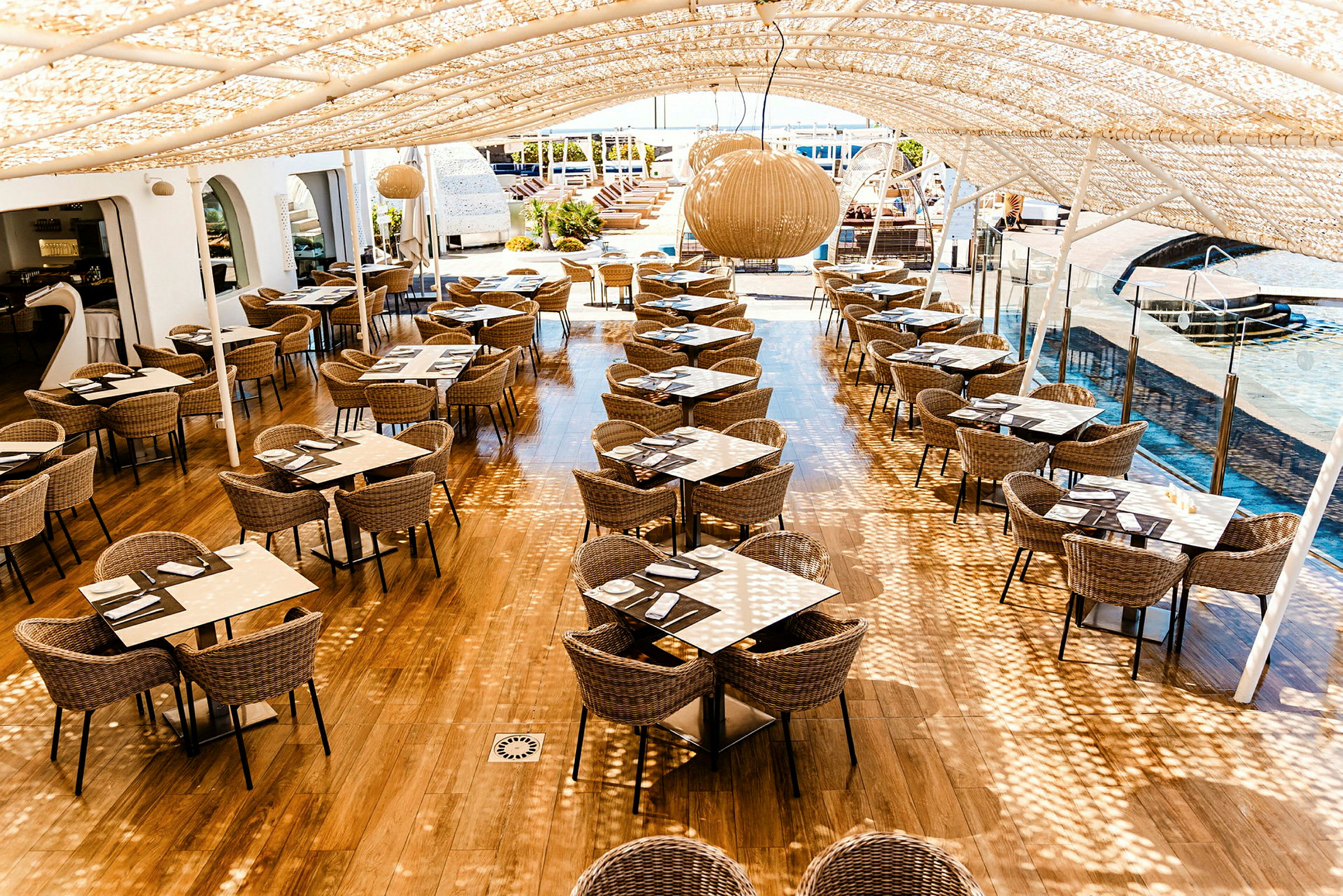 Udendørs restaurant med skyggetag og siddepladser ved borde af kurvestole og glas. (AI-genereret tekst)