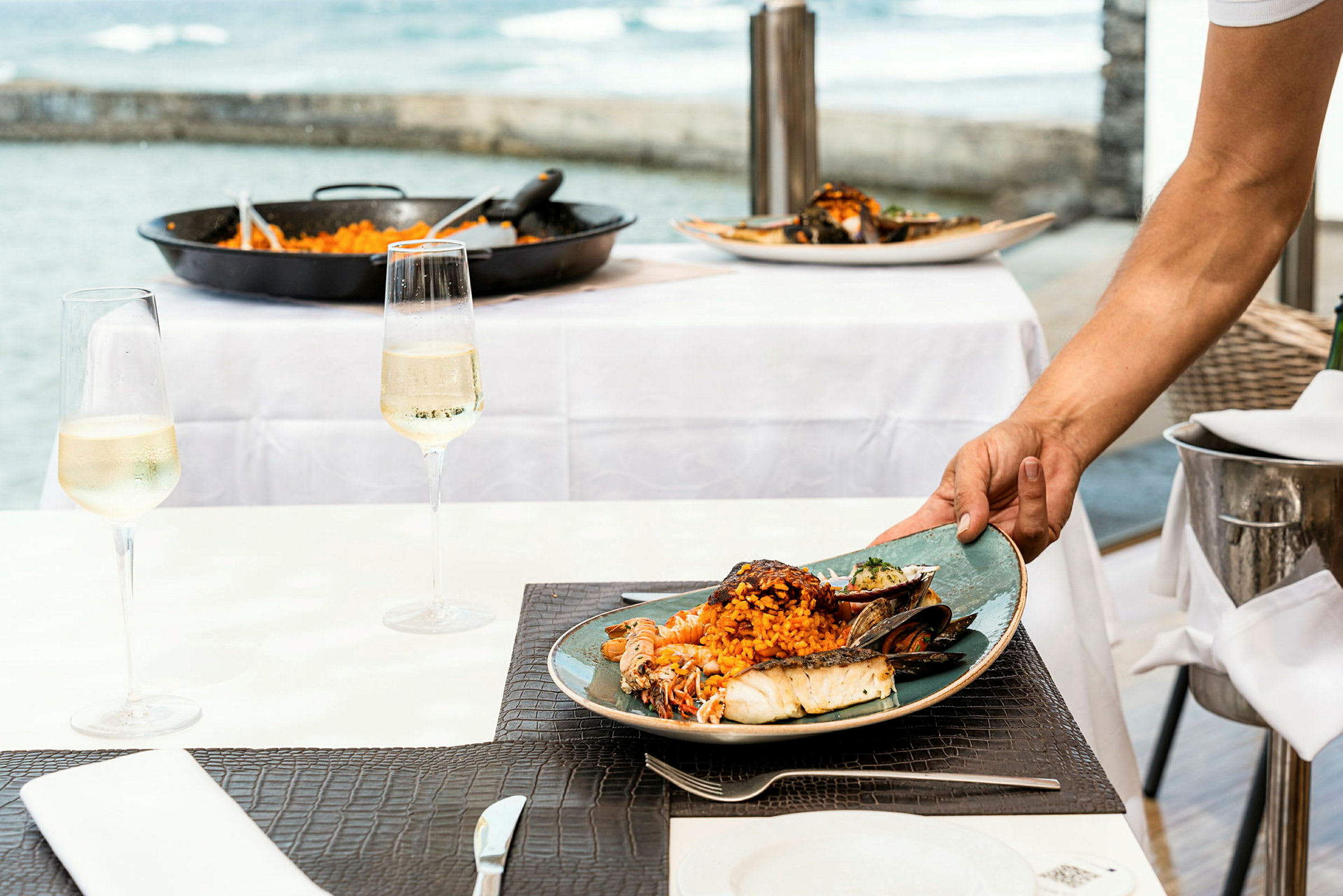 En tallerken med skaldyr og ris serveres ved et bord. En havudsigt ses bag bordet, og et glas vin står på bordet. (AI-genereret tekst)