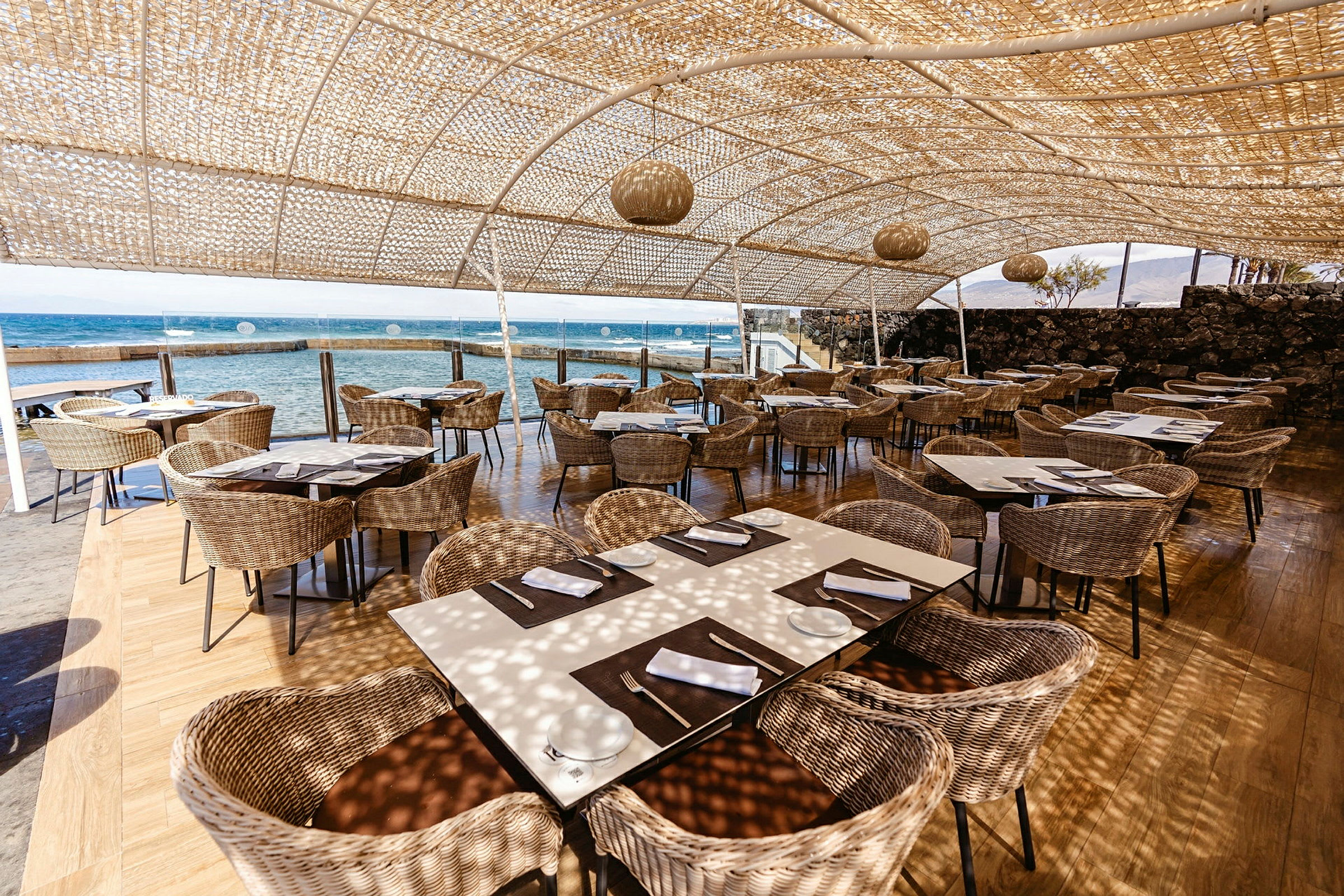 Udendørs restaurant med rottingsæder og udsigt over havet. (AI-genereret tekst)
