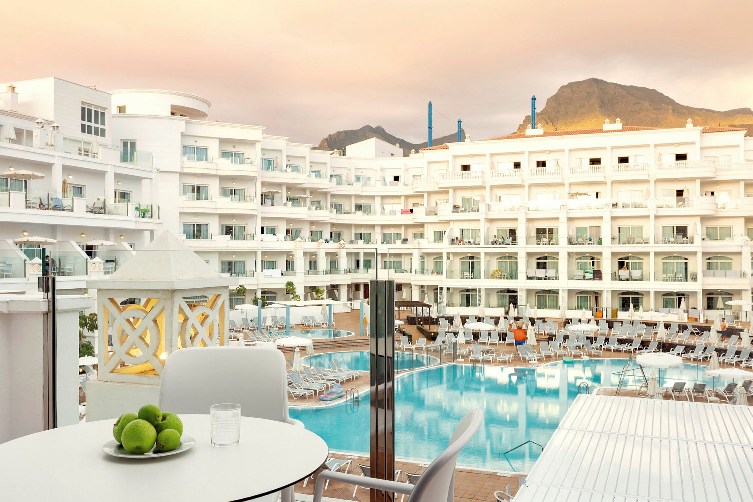 Et hotelkompleks med flere etager, en stor pool og liggestole i forgrunden, samt et bord med frugtskål og glas. (AI-genereret tekst)