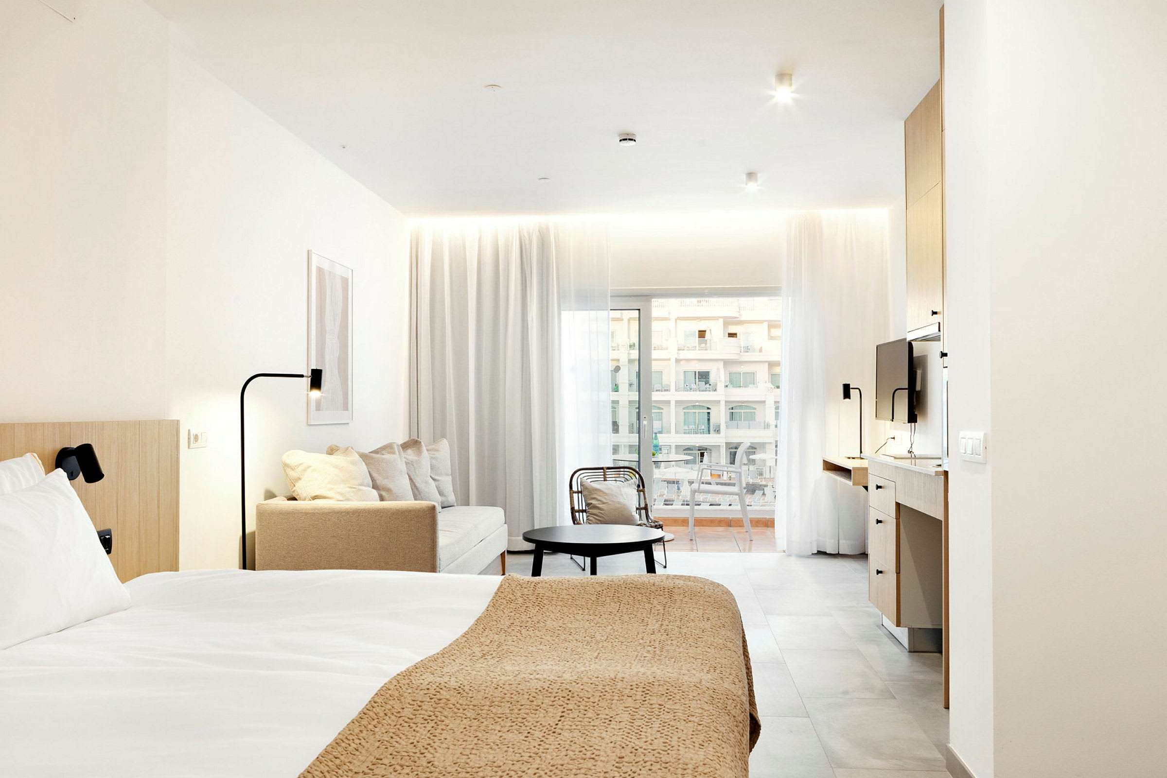 Moderne hotelværelse med seng, siddeområde og arbejdsområde, belyst af naturligt lys fra altandøre. (AI-genereret tekst)