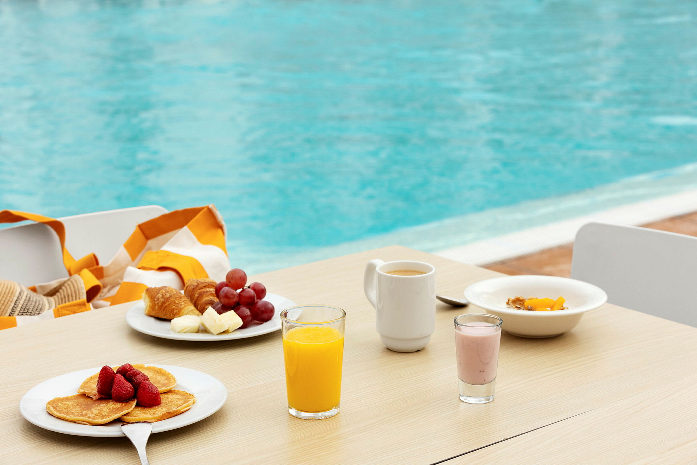 Morgenmad med frugt, pandekager, juice og kaffe på et bord ved en pool. (AI-genereret tekst)