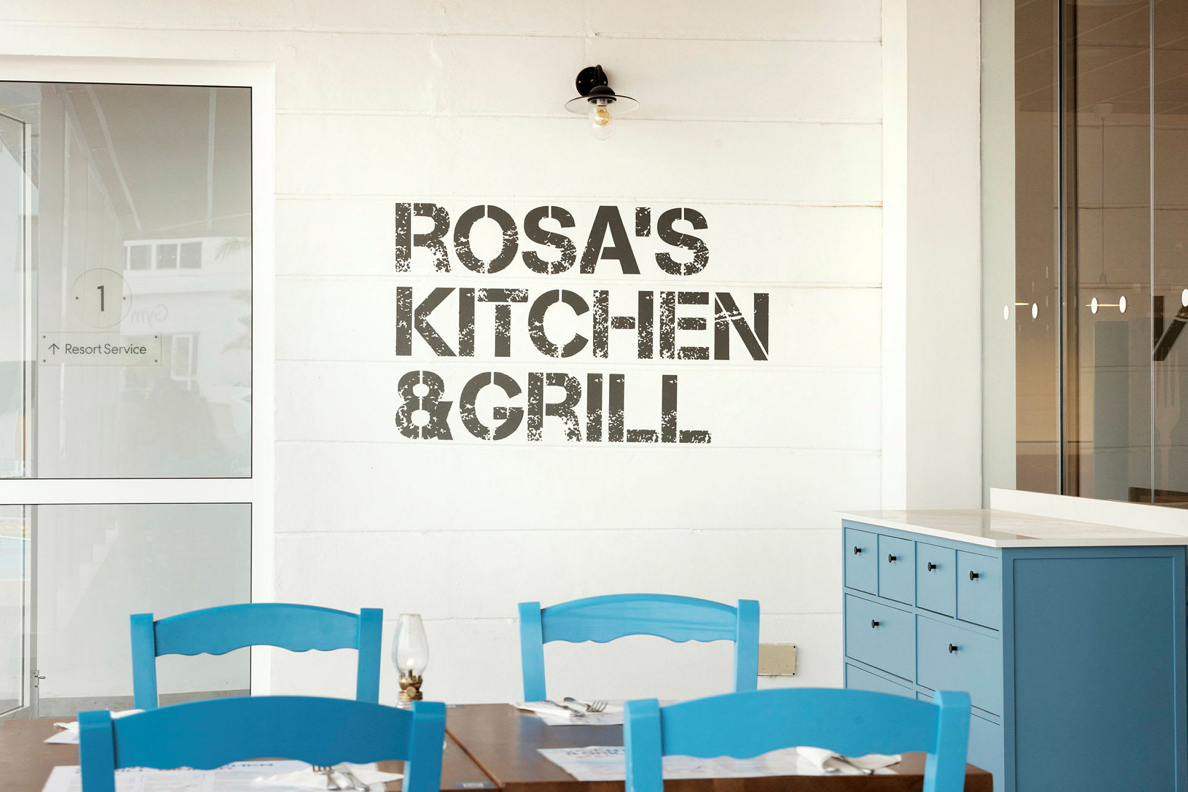 Skilt med teksten 'Rosa's Kitchen & Grill' på en væg, borde og blå stole i forgrunden. (AI-genereret tekst)