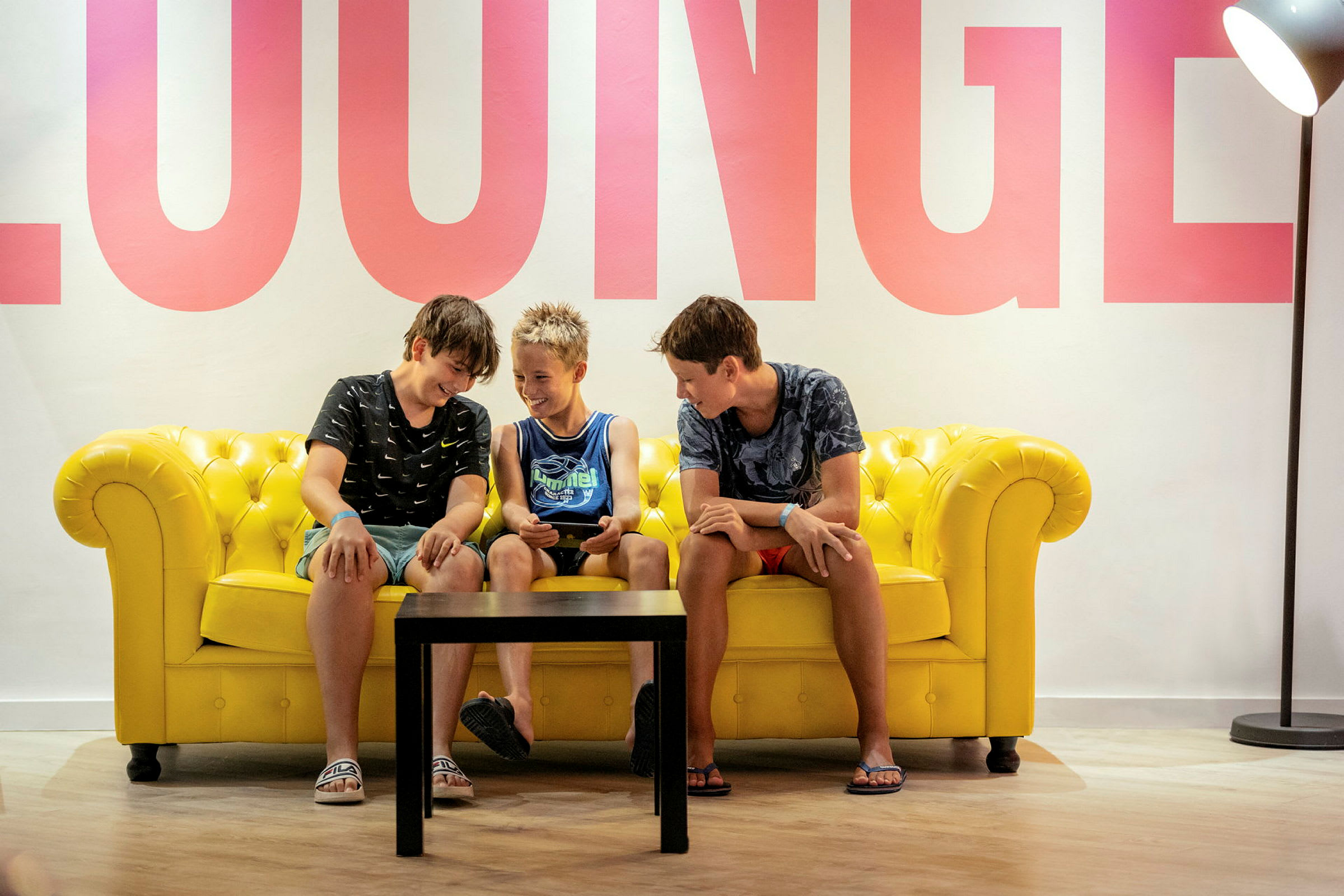 Personer sidder på en gul sofa med et bord foran, og ordet 'LOUNGE' er synligt på væggen. (AI-genereret tekst)