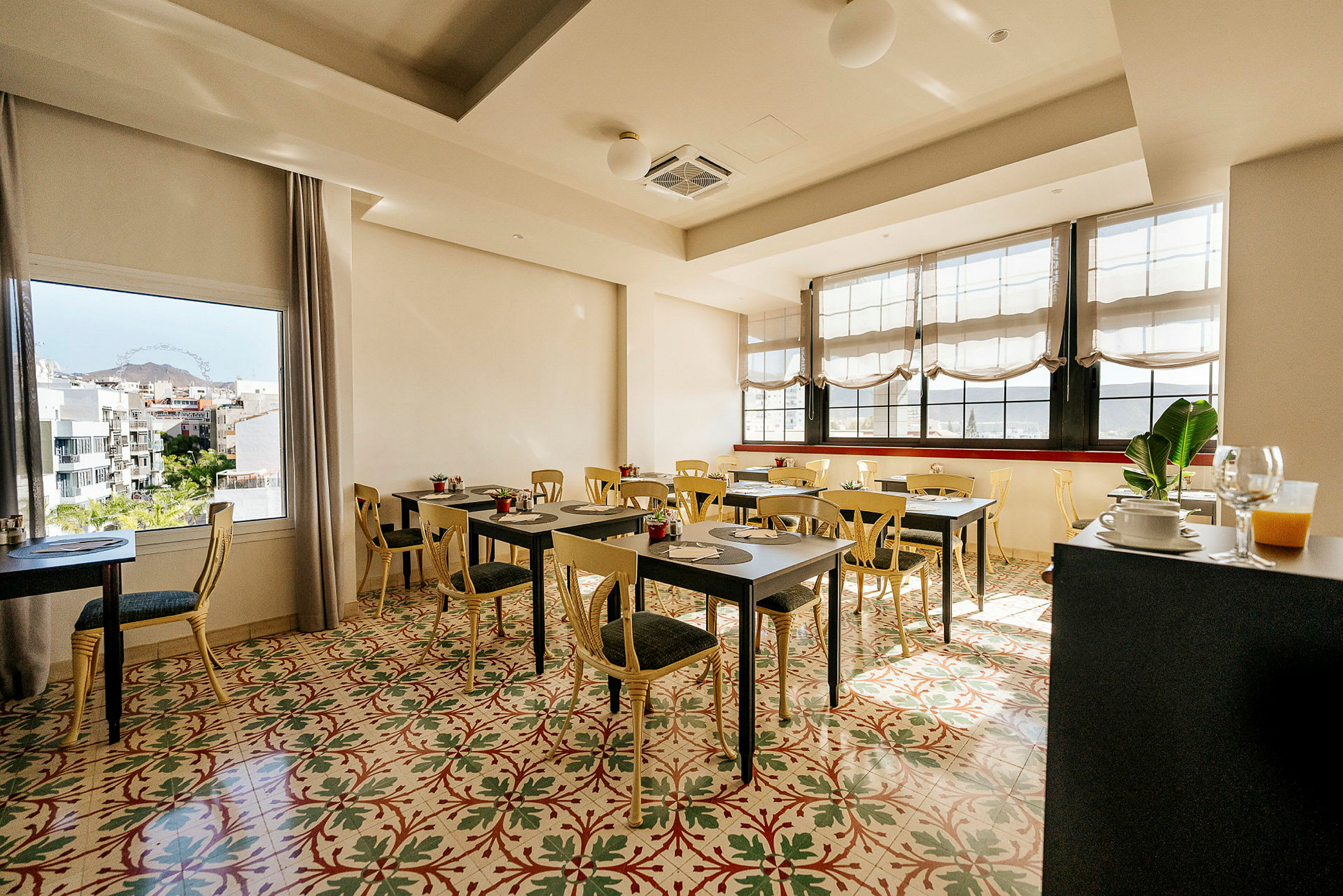 En lys og luftig café eller restaurant med borde og stole, et farverigt gulvmønster, store vinduer og udsigt over et bylandskab. (AI-genereret tekst)