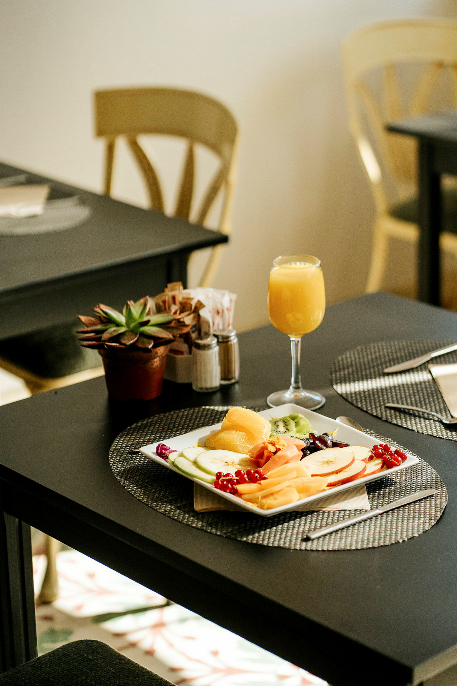 Et bord med en tallerken fyldt med frugt, et glas appelsinjuice og en potteplante. (AI-genereret tekst)