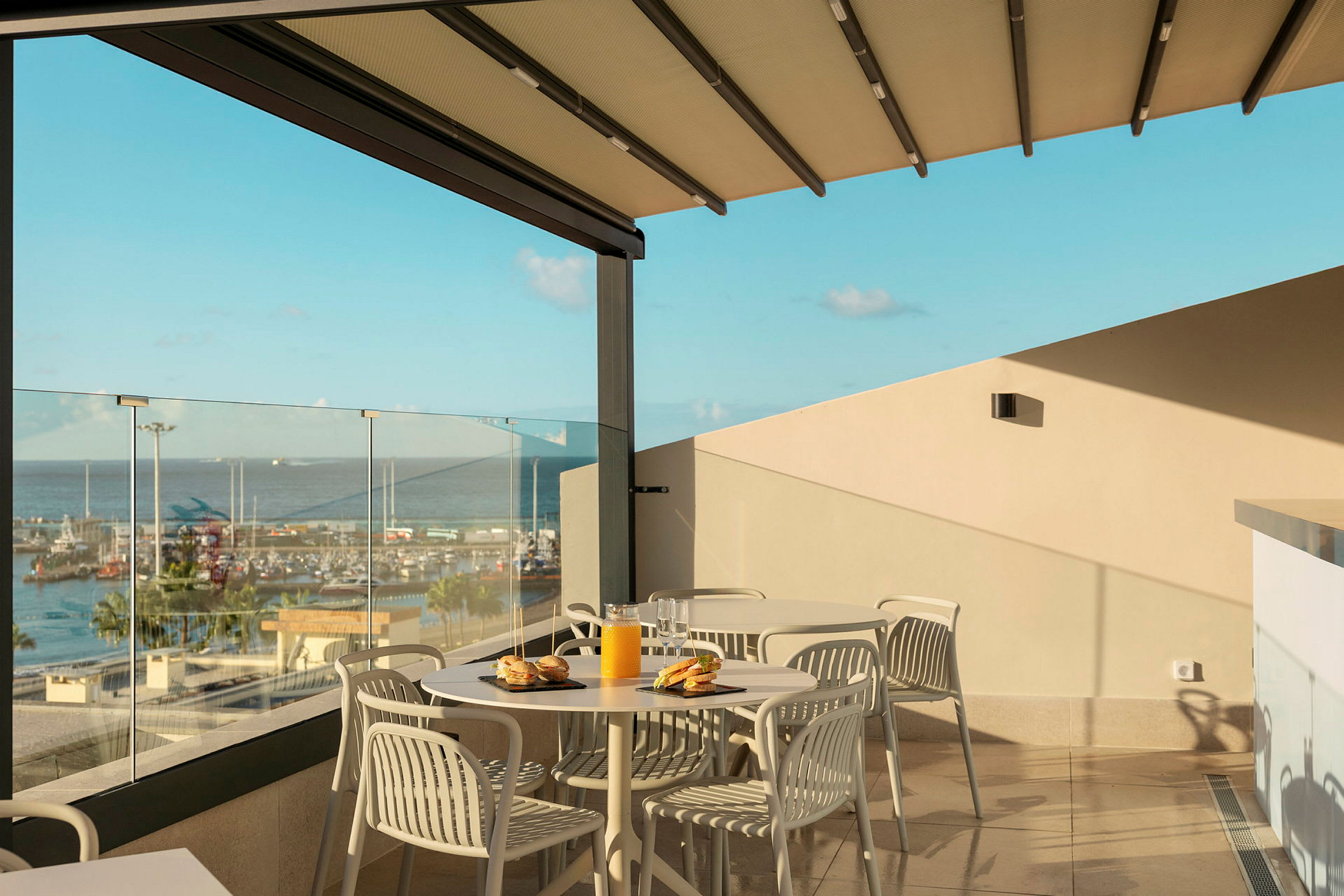 Udsigt fra en terrasse med borde og stole, glasrækværk og udsigt over en havn. (AI-genereret tekst)