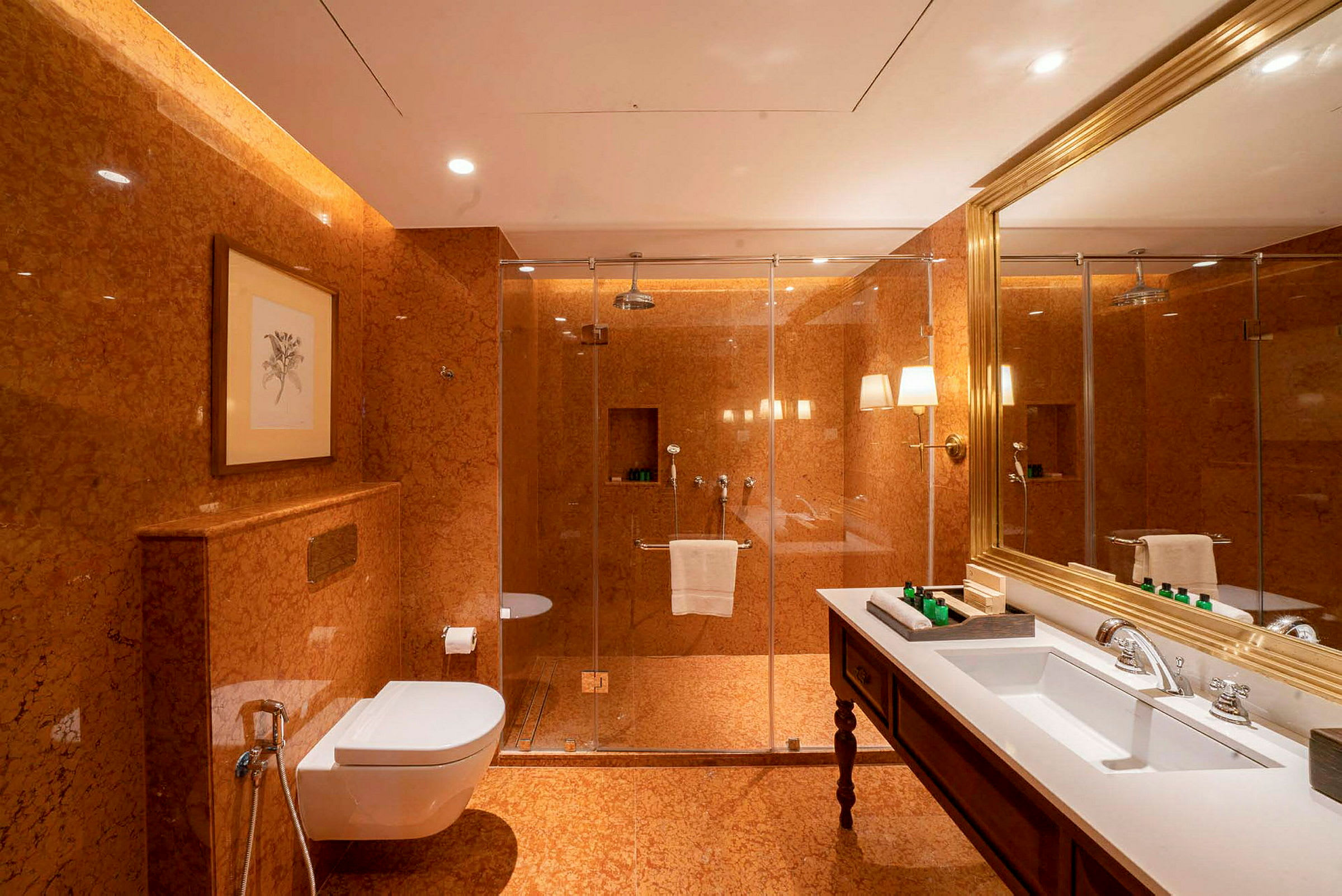Et badeværelse med orange marmorvægge og klinkegulv. Der er en glasbruser, et hvidt toilet med brusehåndtag, et stort spejl med guldramme og et hvidt håndvask med tilhørende hane. (AI-genereret tekst)