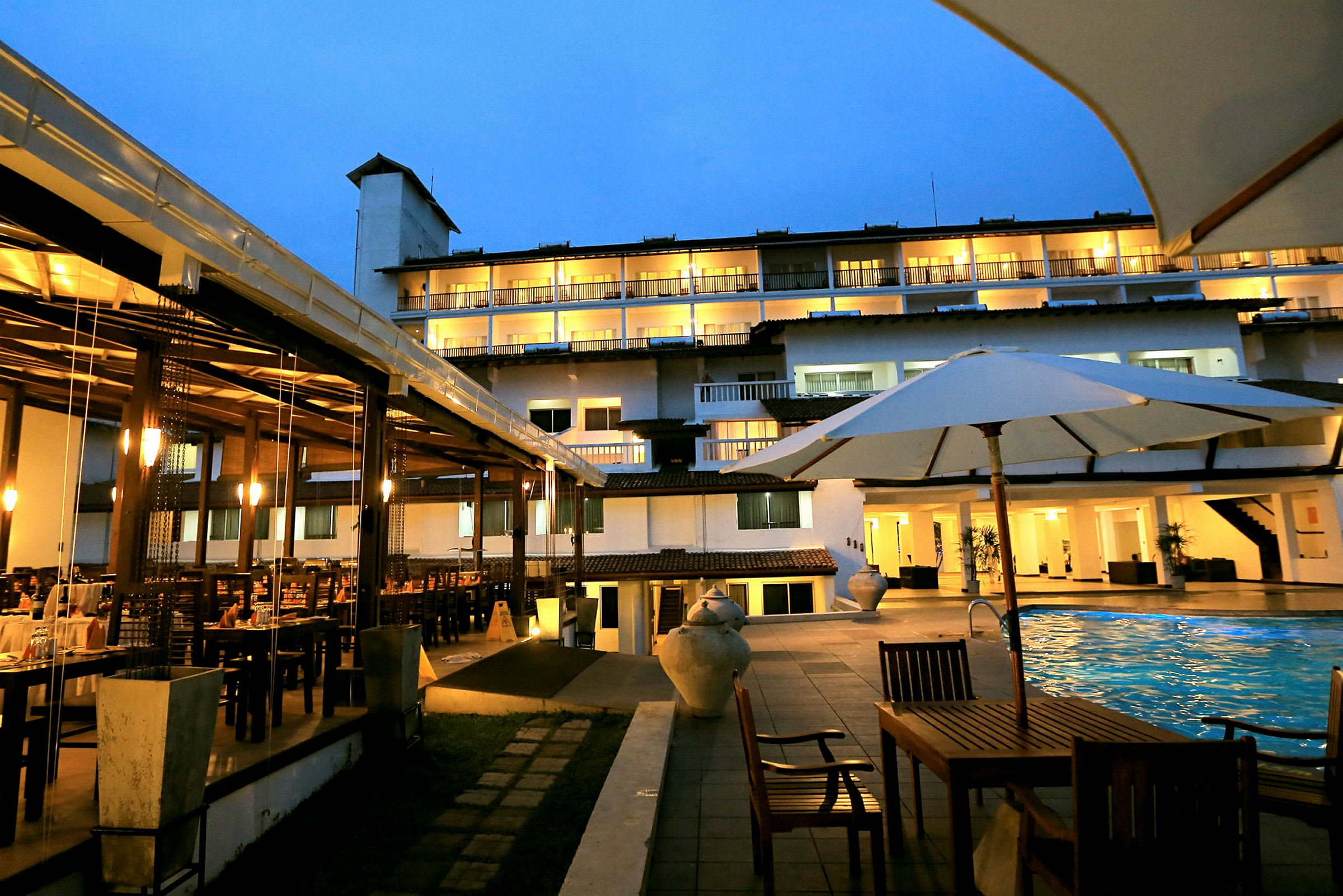 Billede af en hotelbygning om aftenen med oplyste altaner, udendørs pool, havemøbler og et spiseområde. (AI-genereret tekst)