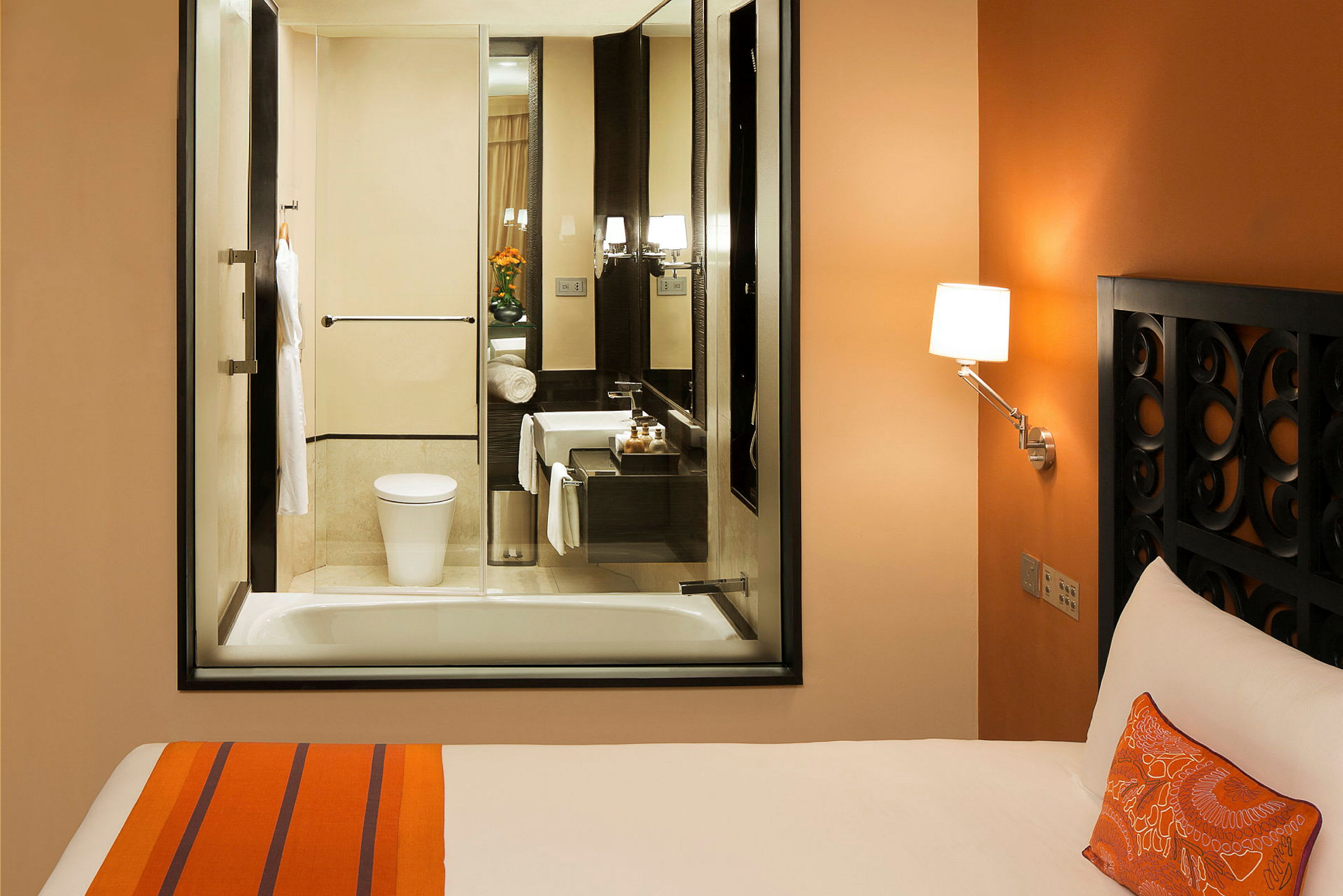 Hotelværelse med orange indretning, seng og et tilstødende badeværelse med glasvindue. (AI-genereret tekst)