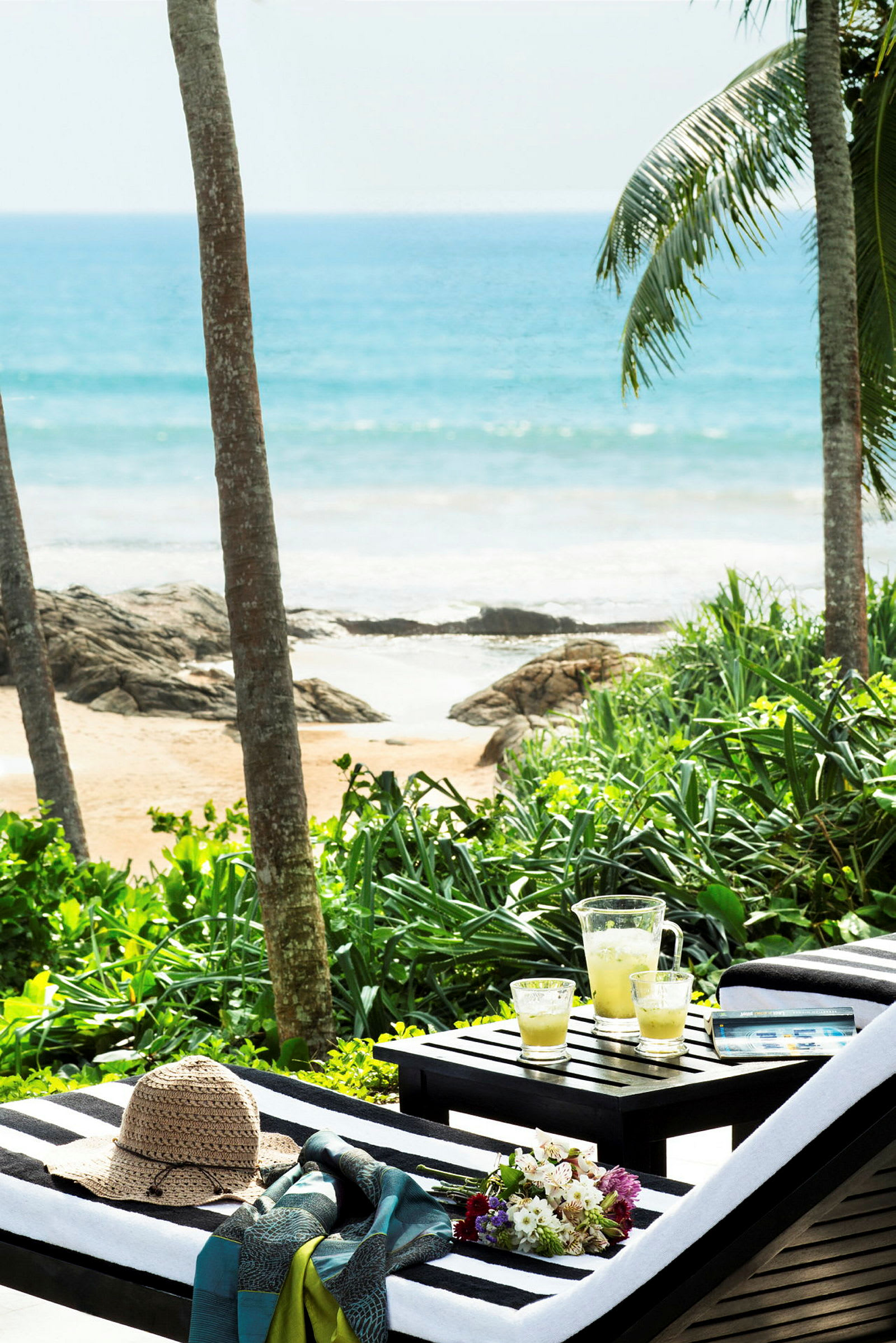 Liggestole ved en tropisk strand med nogle drinks og en hat placeret på bordpladen i forgrunden. (AI-genereret tekst)