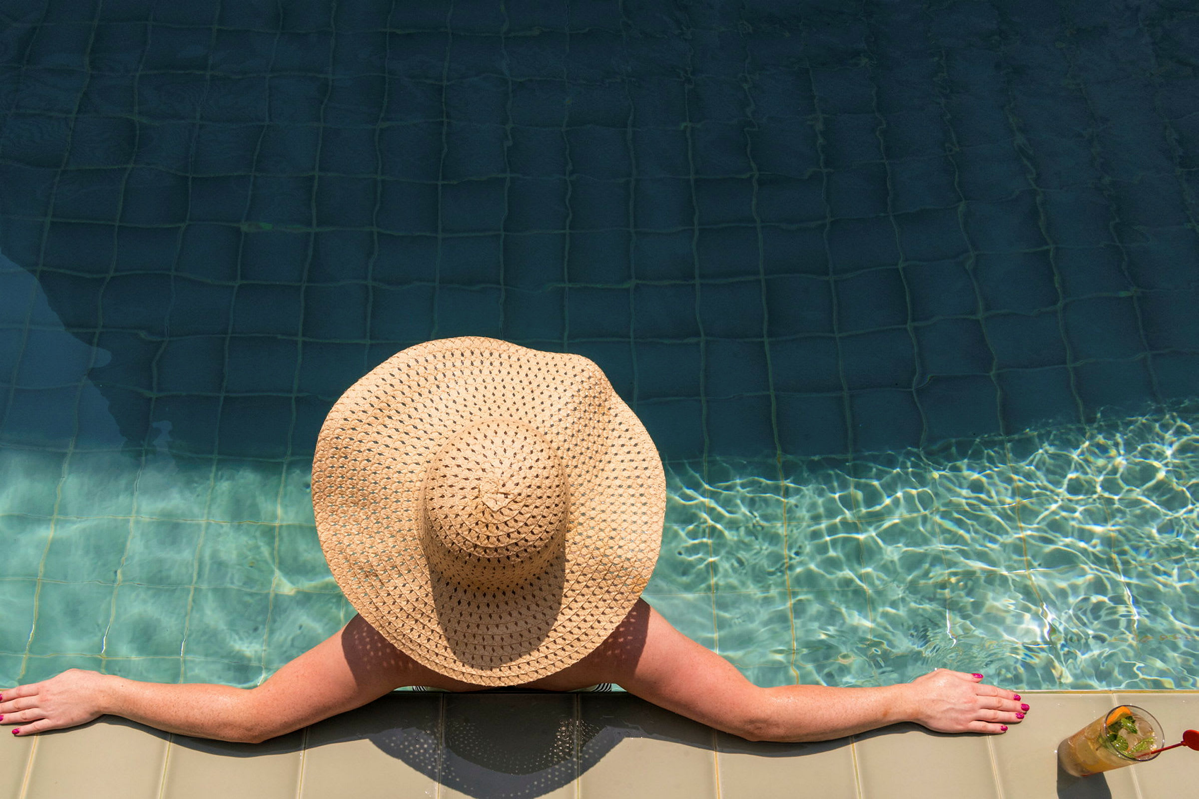 En person iført en bredskygget solhat læner sig mod kanten af en swimmingpool. (AI-genereret tekst)