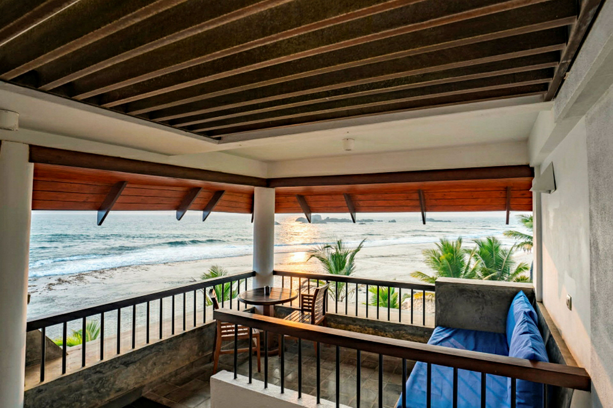 En terrasse med udsigt over stranden og havet. Der er en træstol og et rundt bord samt en siddeplads med en blå pude. (AI-genereret tekst)