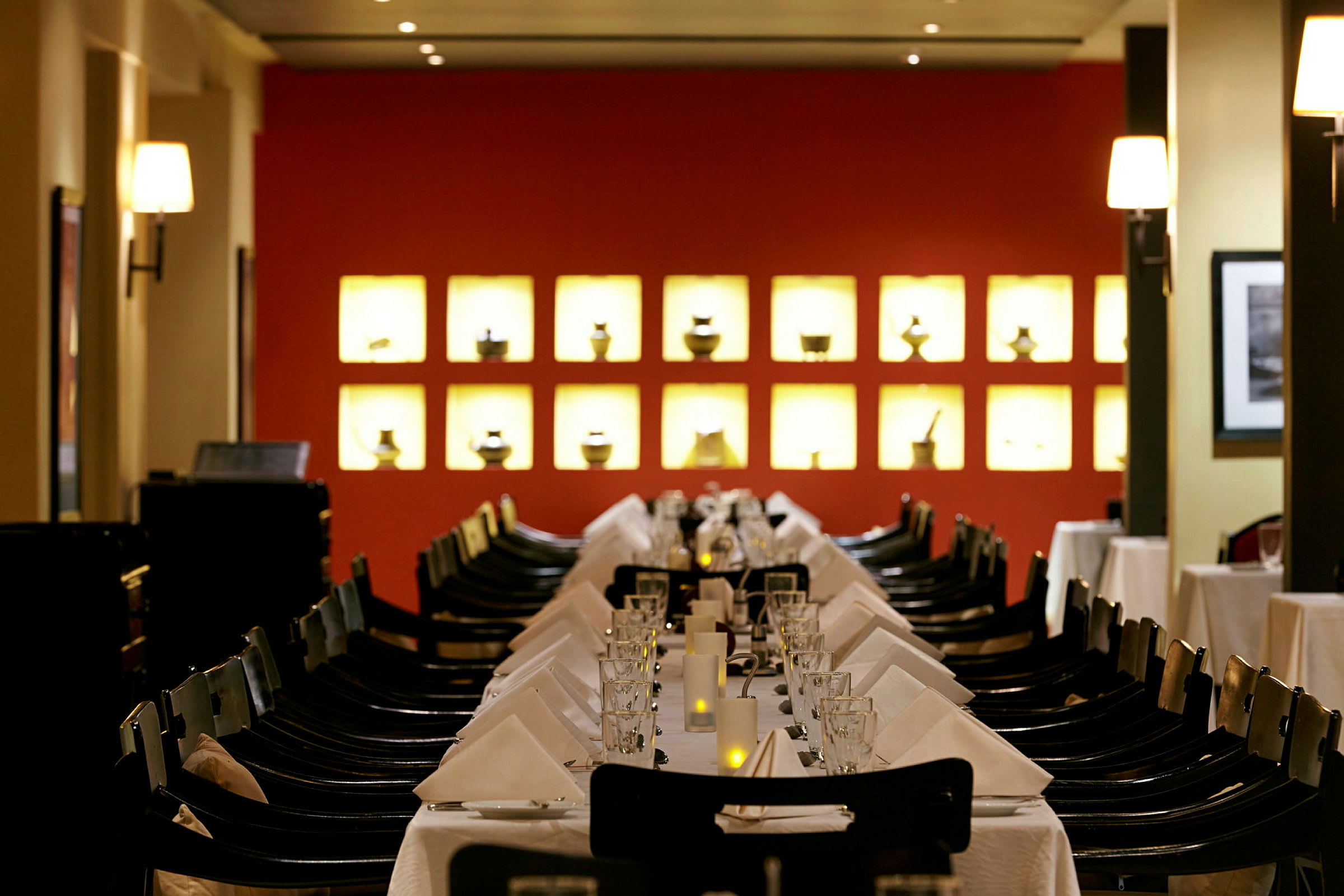 En restaurant med et dækket bord med hvide duge, glas og servietter. I baggrunden er der en rød væg med oplyste små dekorative genstande. (AI-genereret tekst)