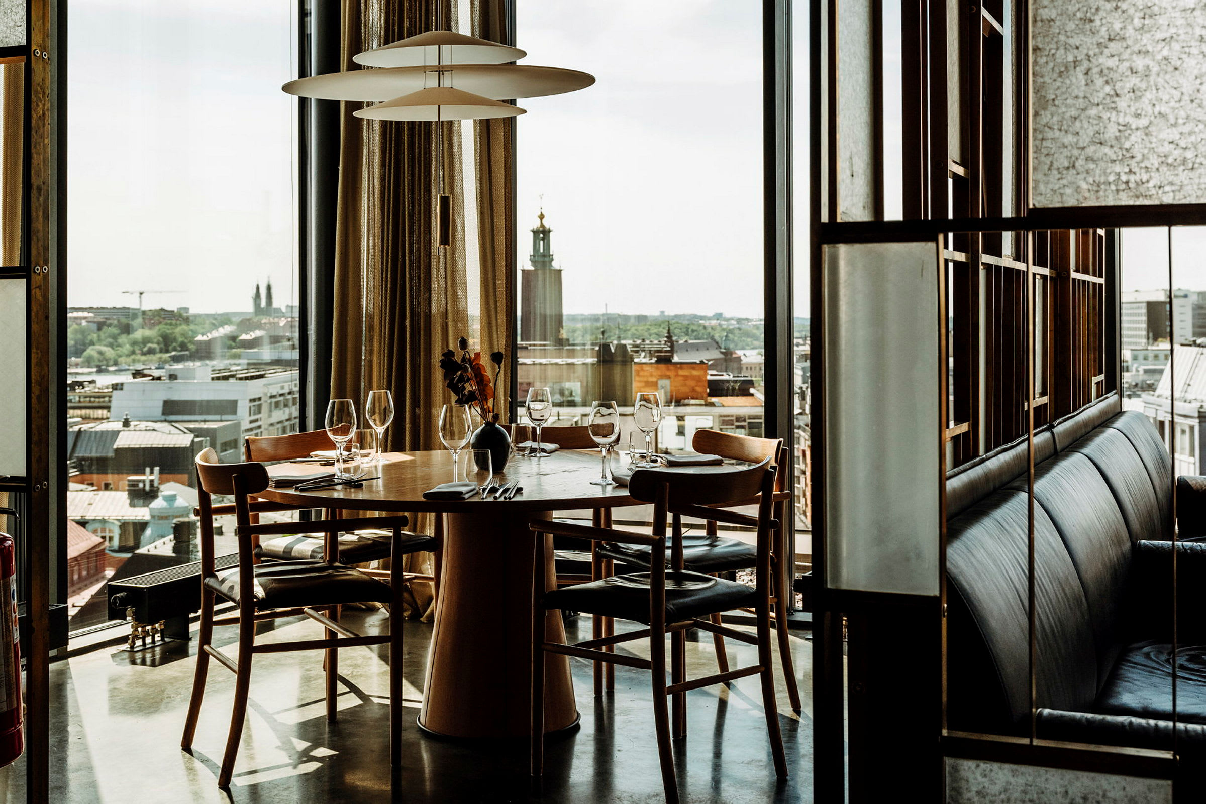 Et dækket bord til fire personer i en restaurant med udsigt over en by gennem store vinduer. (AI-genereret tekst)