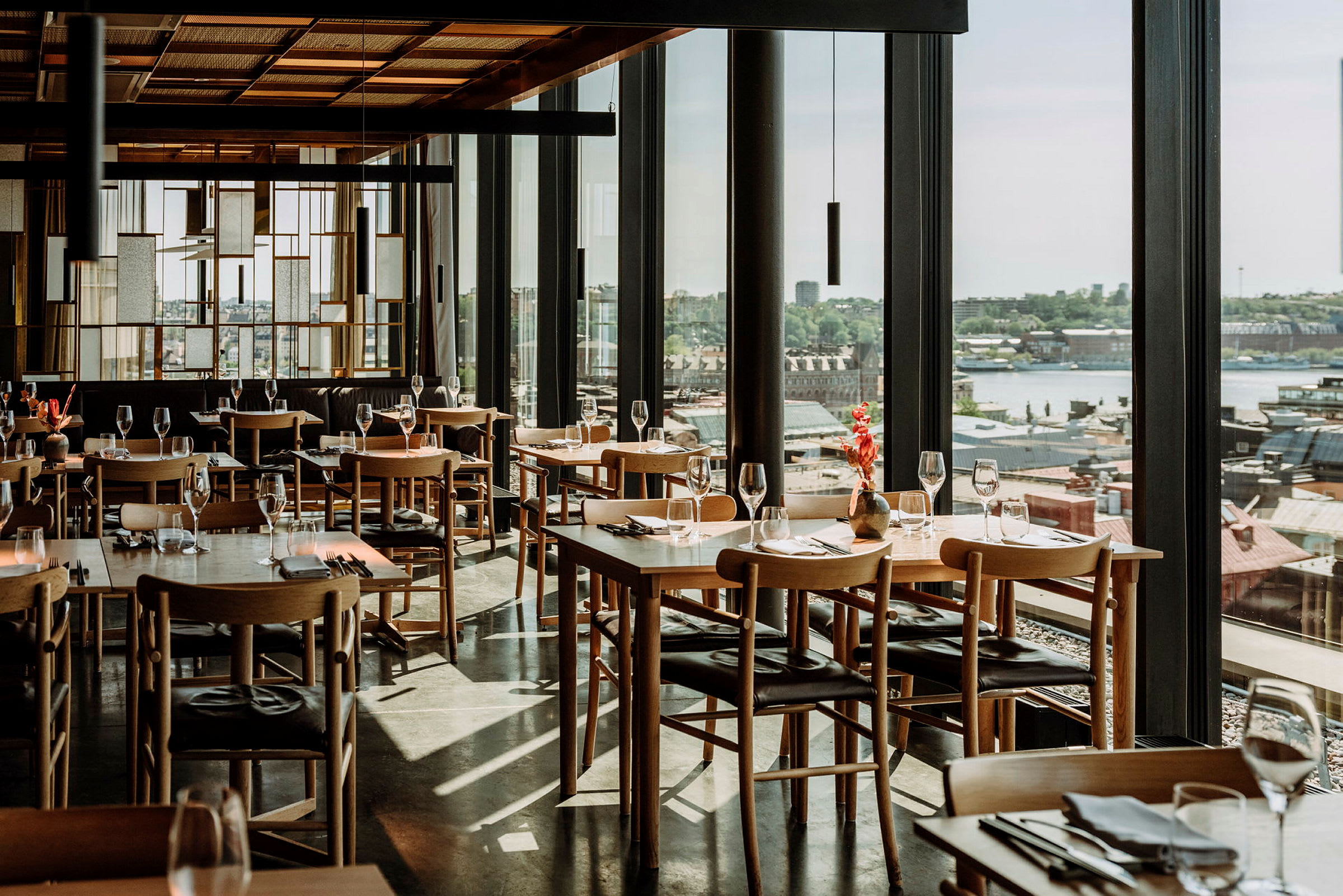 En stilfuld restaurant med træborde og stole, glas der reflekterer lys, og en panoramaudsigt over en by. (AI-genereret tekst)
