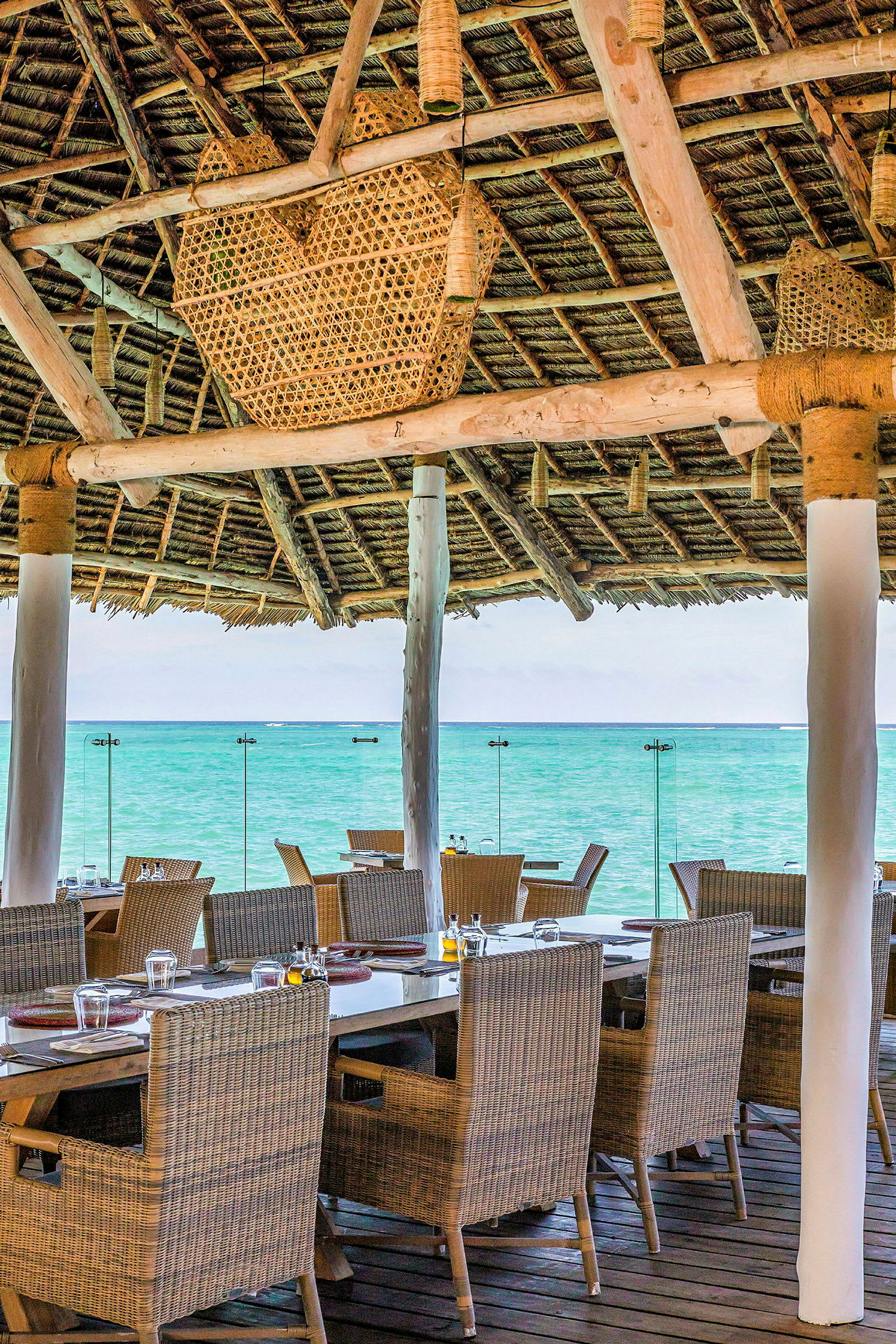 Restaurantens siddepladser ved havet med borde og stole under et træ- og stråtag. (AI-genereret tekst)